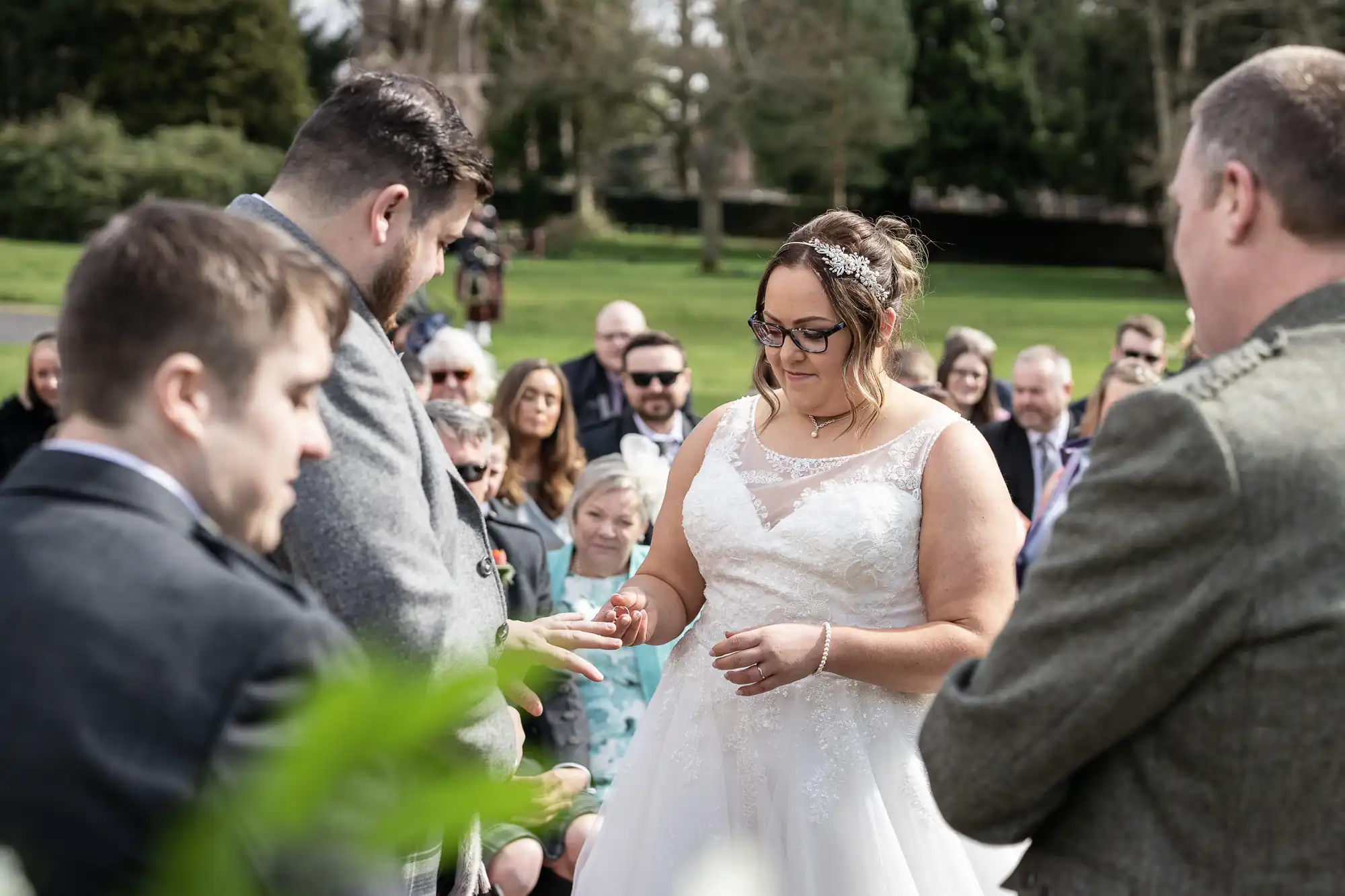 A bride in a white dress places a ring on the groom's finger during an outdoor wedding ceremony. Guests, dressed semi-formally, watch from their seats. Trees and greenery fill the background, and two men stand beside the couple.