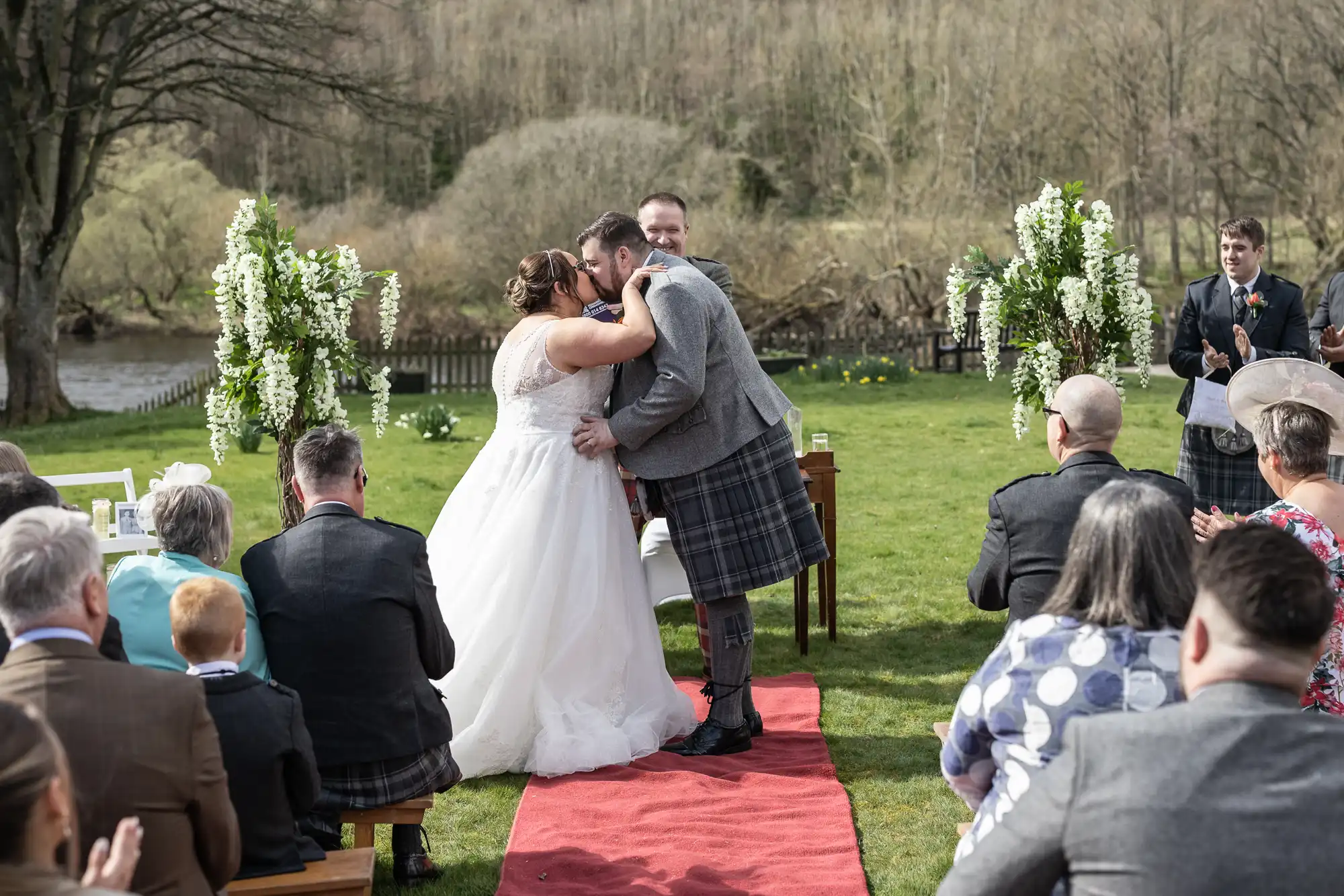 A bride and groom kiss at an outdoor wedding ceremony on a grassy lawn, surrounded by guests in formal attire. The guests are seated facing the couple, and tall flower arrangements decorate the aisle. Trees and a river are visible in the background.