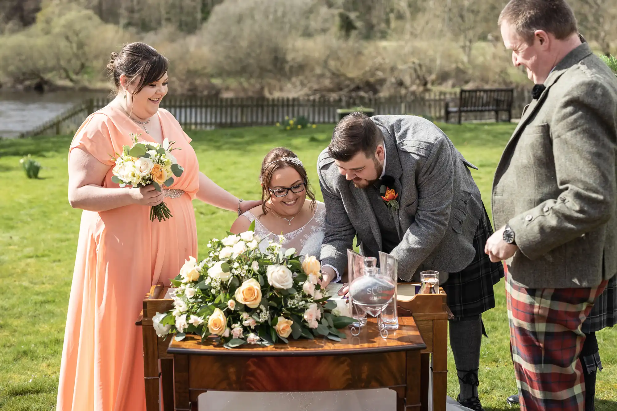 Three people stand around a seated woman signing a document on a wooden table decorated with cream and peach flowers. The scene is outdoors on grass, with trees and a river in the background. All are dressed formally, suggesting a wedding ceremony.