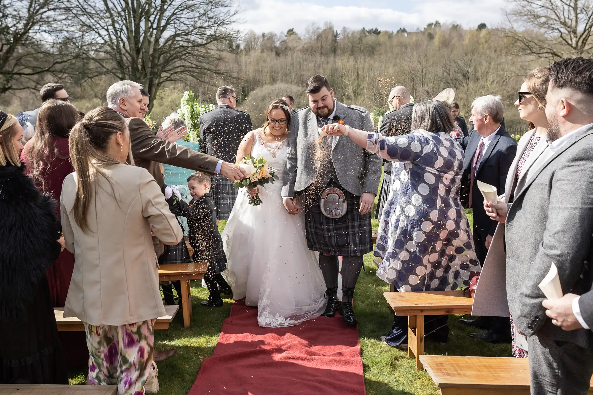 A bride in a white gown and a groom in a plaid kilt walk down a red aisle outdoors, smiling as guests on both sides toss confetti. People are dressed in formal attire, and trees and cloudy skies are visible in the background.