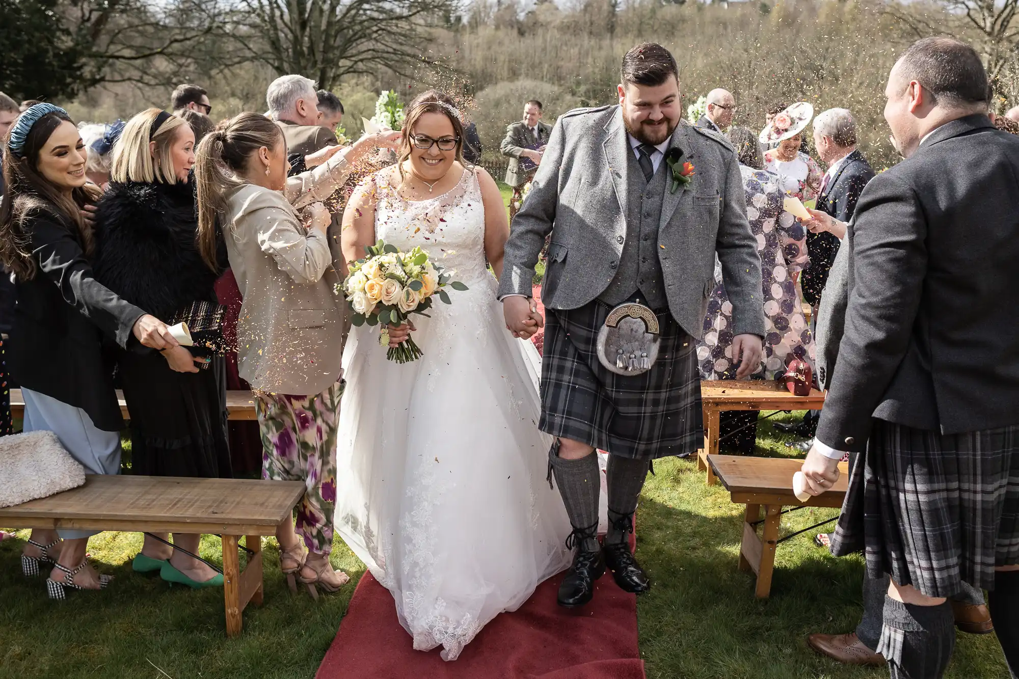 A smiling bride in a white gown and a groom in a grey jacket and kilt walk hand in hand down a red aisle outdoors, as guests cheer and throw confetti. The scene is joyful, with greenery in the background and people dressed festively.