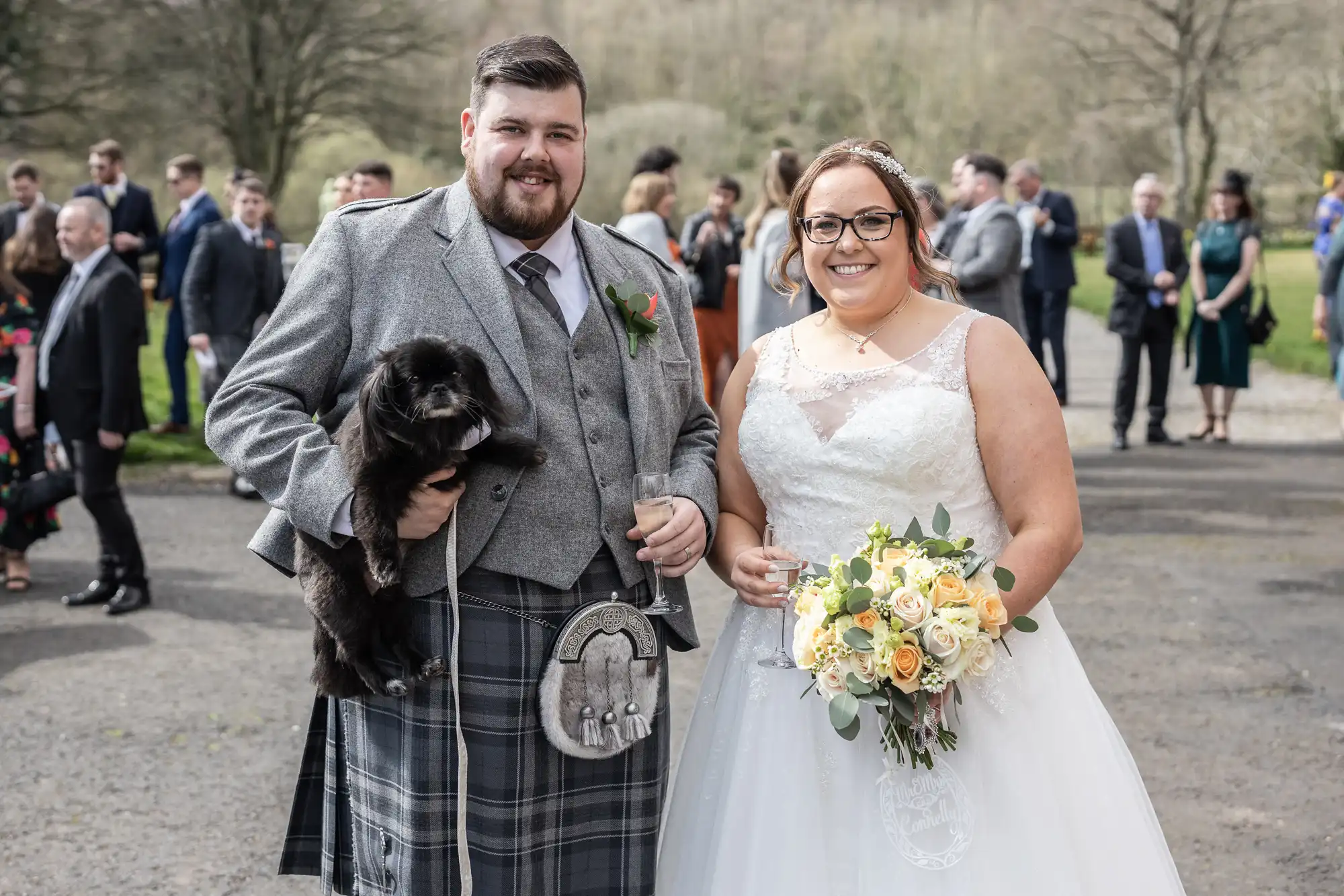 A smiling bride in a white gown holds a bouquet of yellow and white flowers next to a groom in a grey jacket, kilt, and sporran, holding a black dog. Wedding guests stand blurred in the background outdoors on a cloudy day.