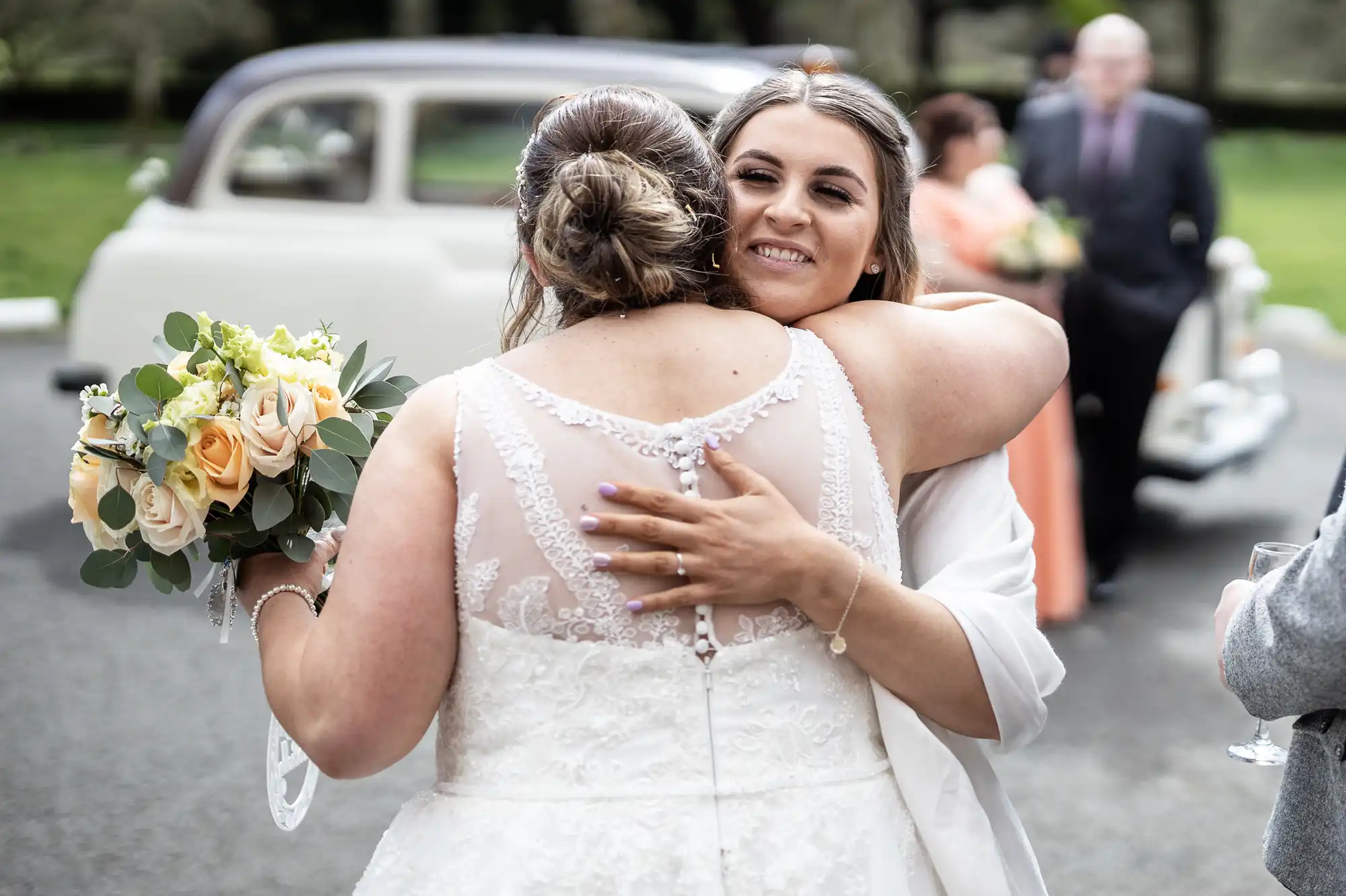 A bride in a lace wedding dress hugs a smiling woman outdoors. The bride holds a bouquet of yellow roses. Classic car and guests in formal attire are blurred in the background. The moment captures joy and celebration.