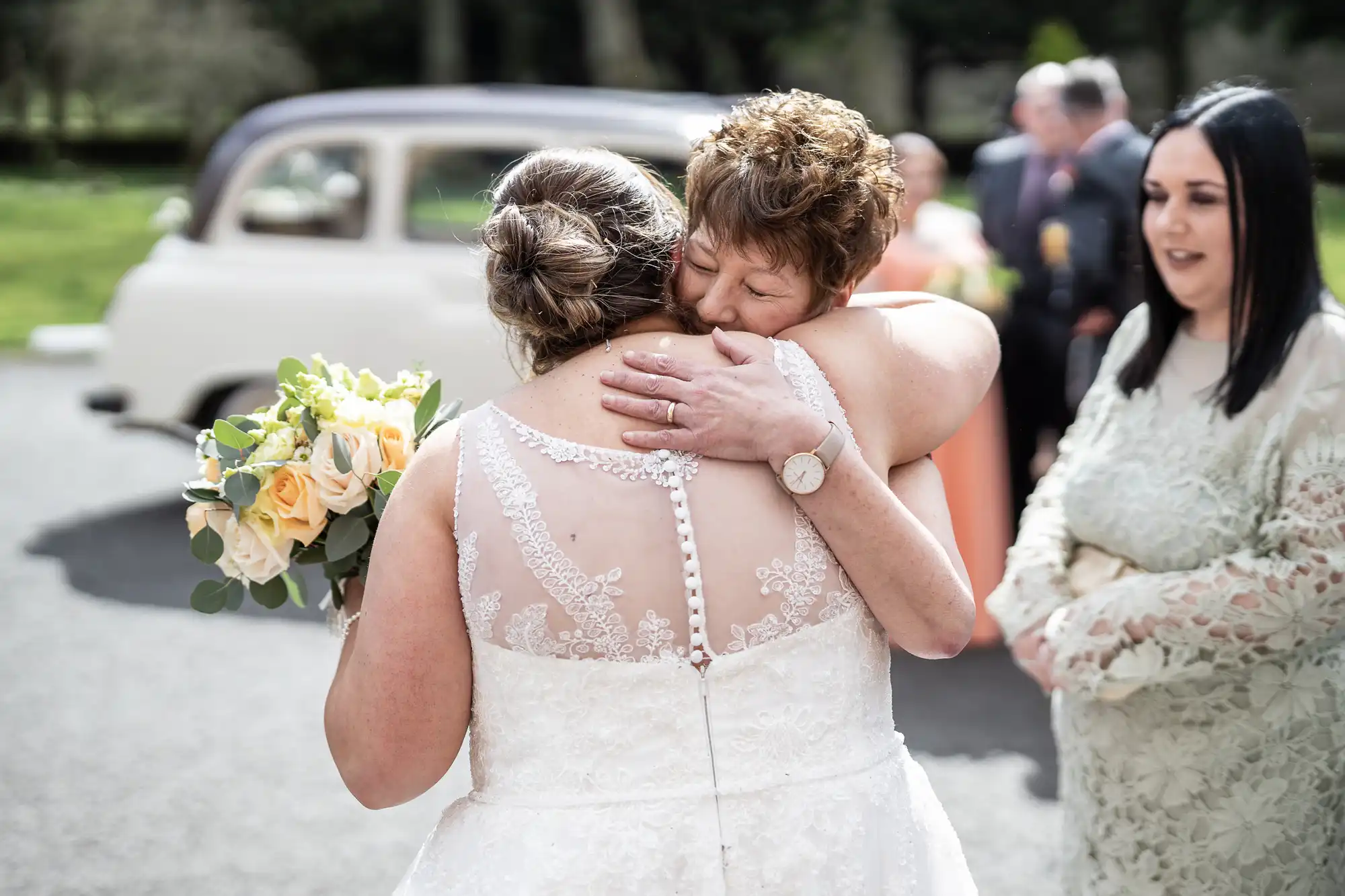 A bride in a white lace dress hugs an older woman outside, her bouquet of yellow roses in hand. The older woman closes her eyes, smiling warmly. People and a vintage car are blurred in the background. Another woman in a pale green dress stands nearby, watching.