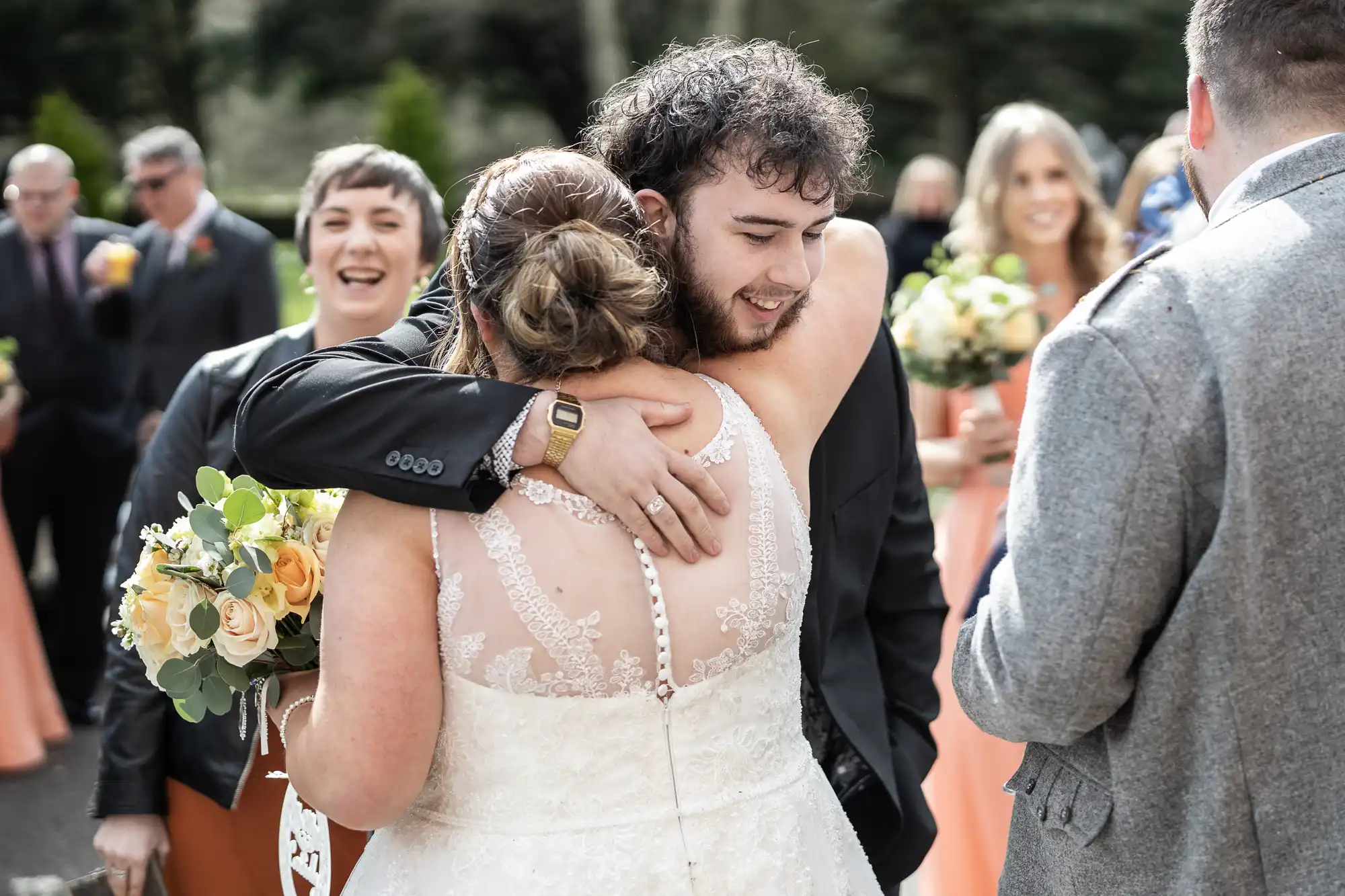 A bride in a lace wedding dress hugs a man in a black suit outdoors, both smiling. Bridesmaids in peach dresses holding bouquets stand nearby, and guests in suits mingle in the background on a sunny day.
