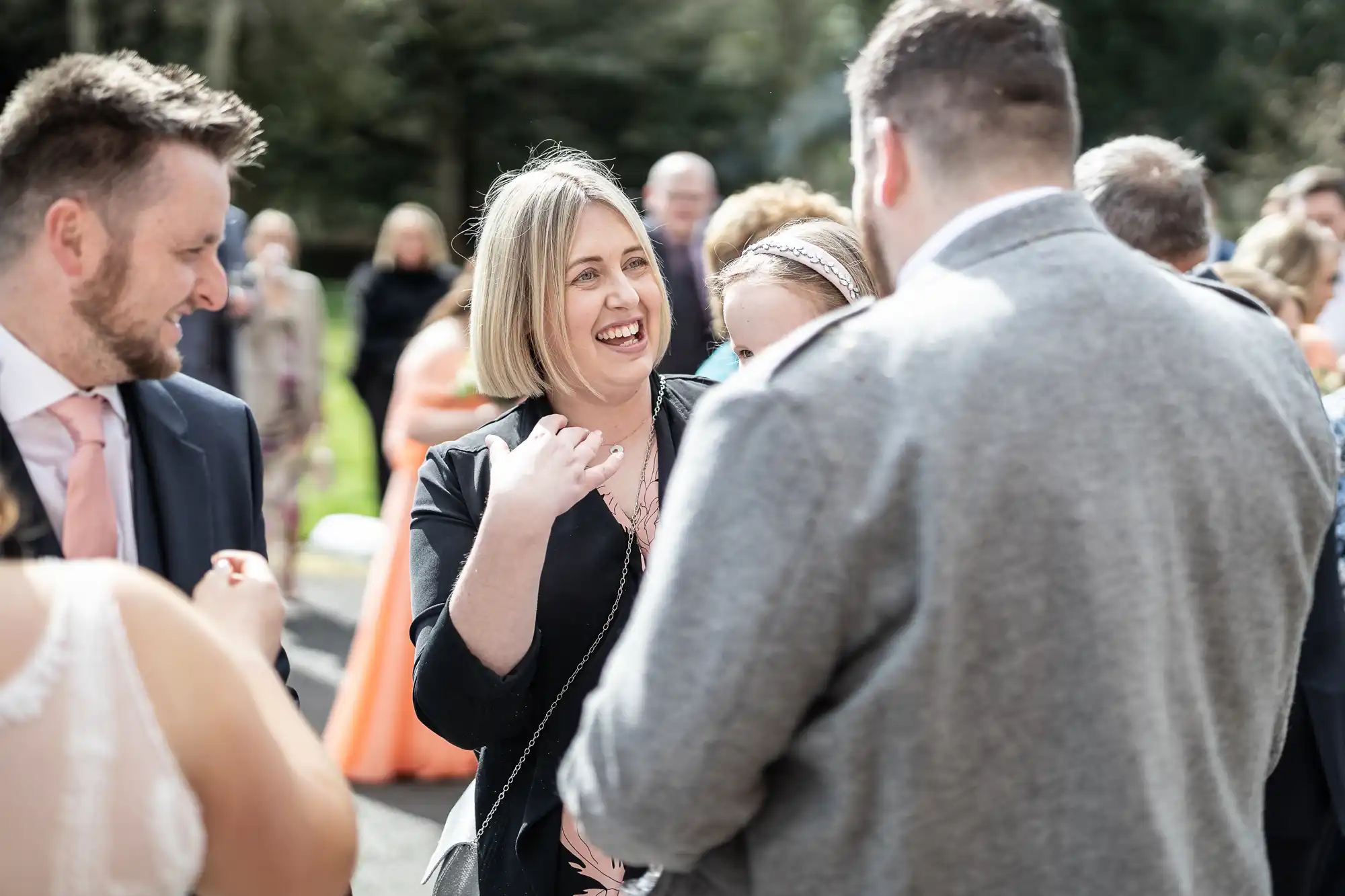 A woman with blonde hair, smiling and wearing a black jacket, stands outdoors among a group of people dressed in formal attire, engaging in conversation at what appears to be a wedding or social event. Sunlight and greenery are visible in the background.