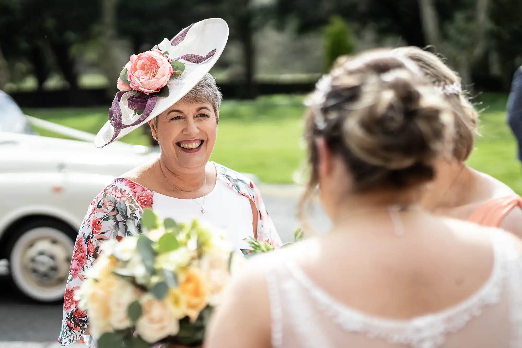 A smiling older woman in a floral hat and shawl stands outdoors, facing two women in light-coloured dresses, one holding a bouquet of yellow roses. A classic white car and green lawn appear blurred in the background.