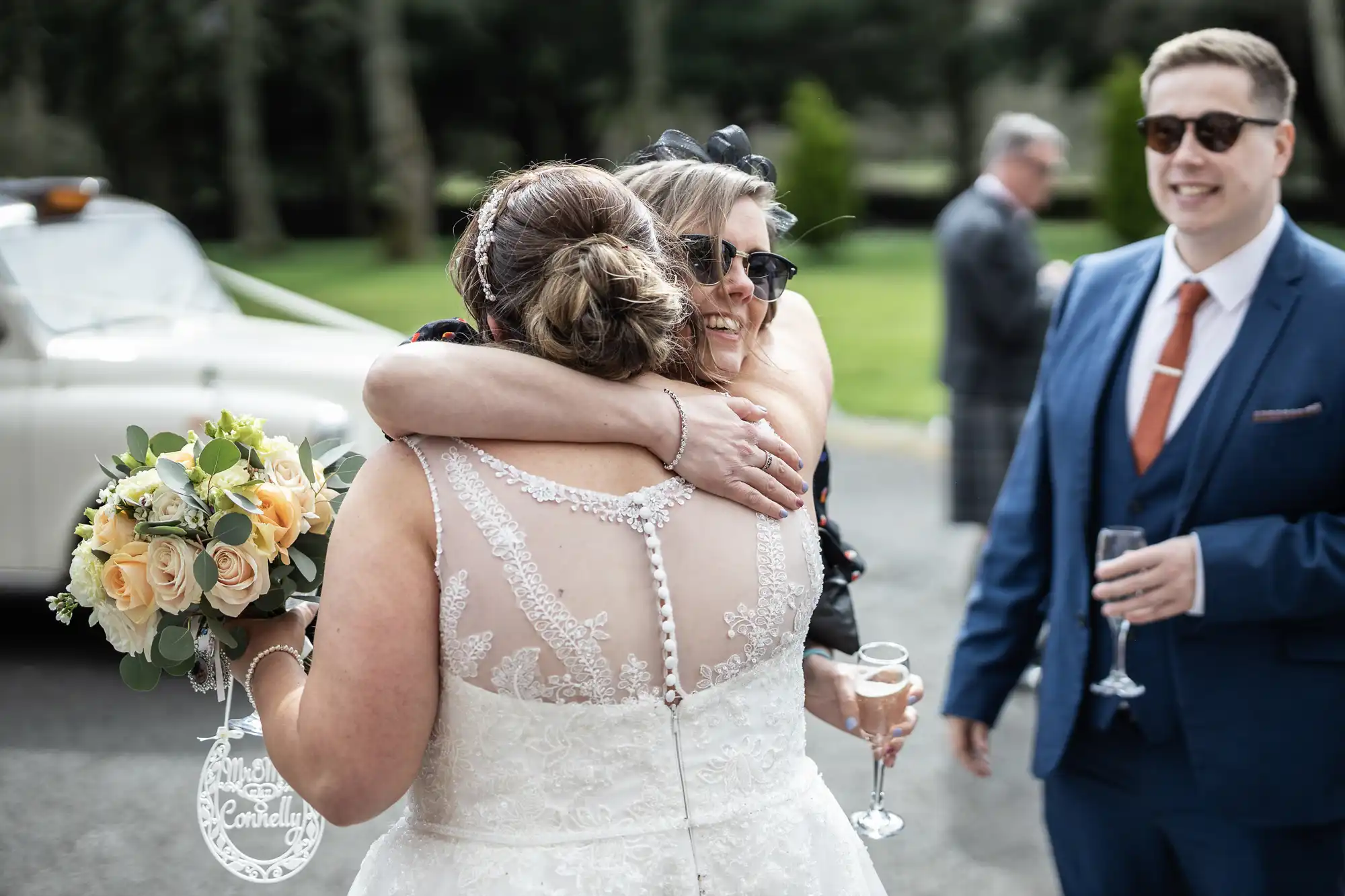 A bride in a white lace dress hugs a smiling woman wearing sunglasses. The bride holds a bouquet of peach and white flowers. A man in a blue suit with sunglasses stands nearby, holding a glass. A vintage car and greenery are visible in the background.