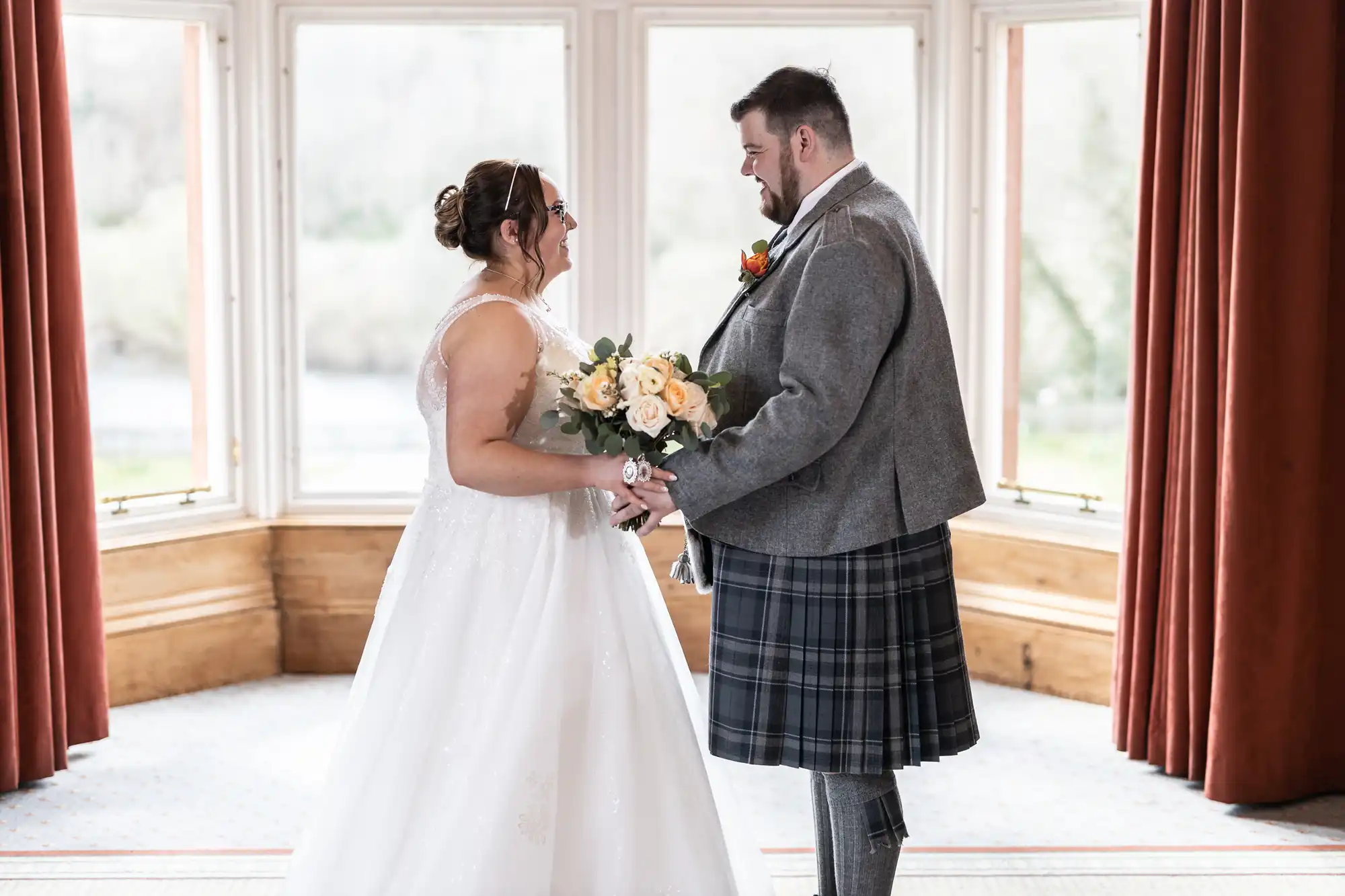 A bride in a white gown holding a bouquet stands facing a groom in a grey jacket and tartan kilt. They hold hands and smile at each other in front of large windows framed by red curtains. Natural light fills the elegant room.