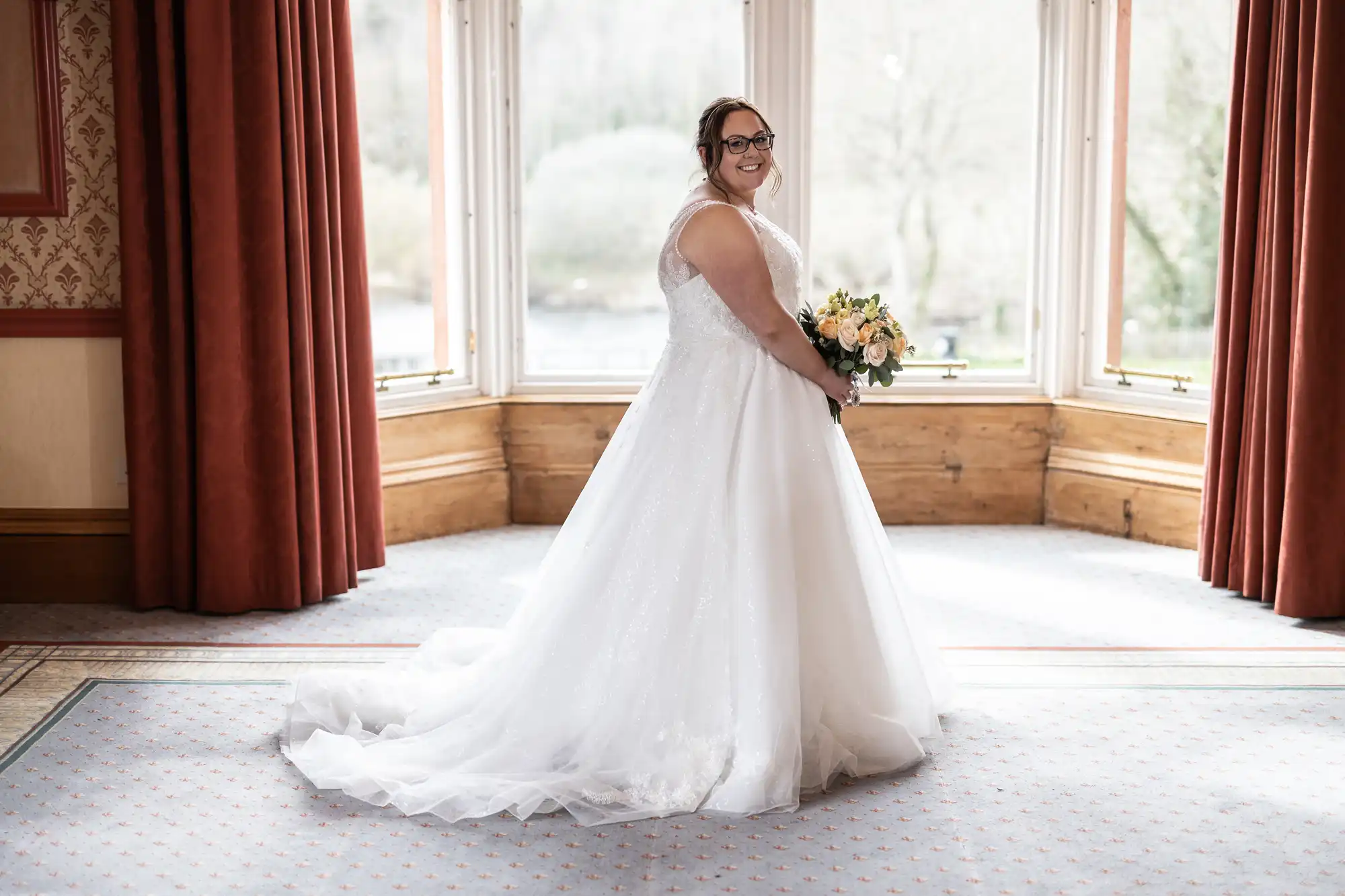A bride in a white gown stands smiling, holding a bouquet of yellow and white flowers. She faces left near large bay windows with red curtains, natural light filling the elegant room. Her dress has a long train and delicate details, and she wears glasses.