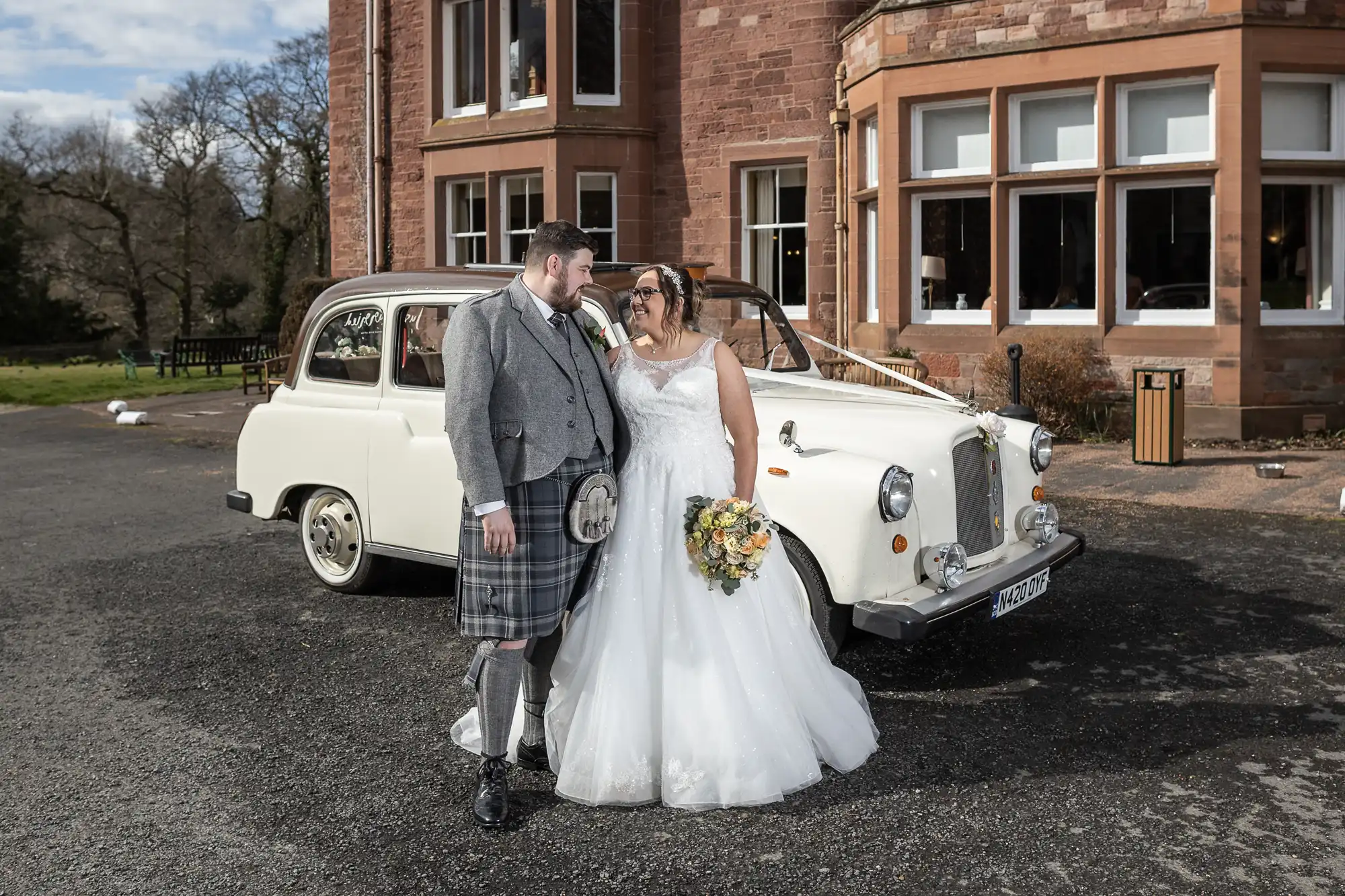 A bride in a white gown and a groom in a grey jacket and kilt smile at each other in front of a vintage white car, with a large stone building and trees in the background. The bride holds a bouquet of flowers.