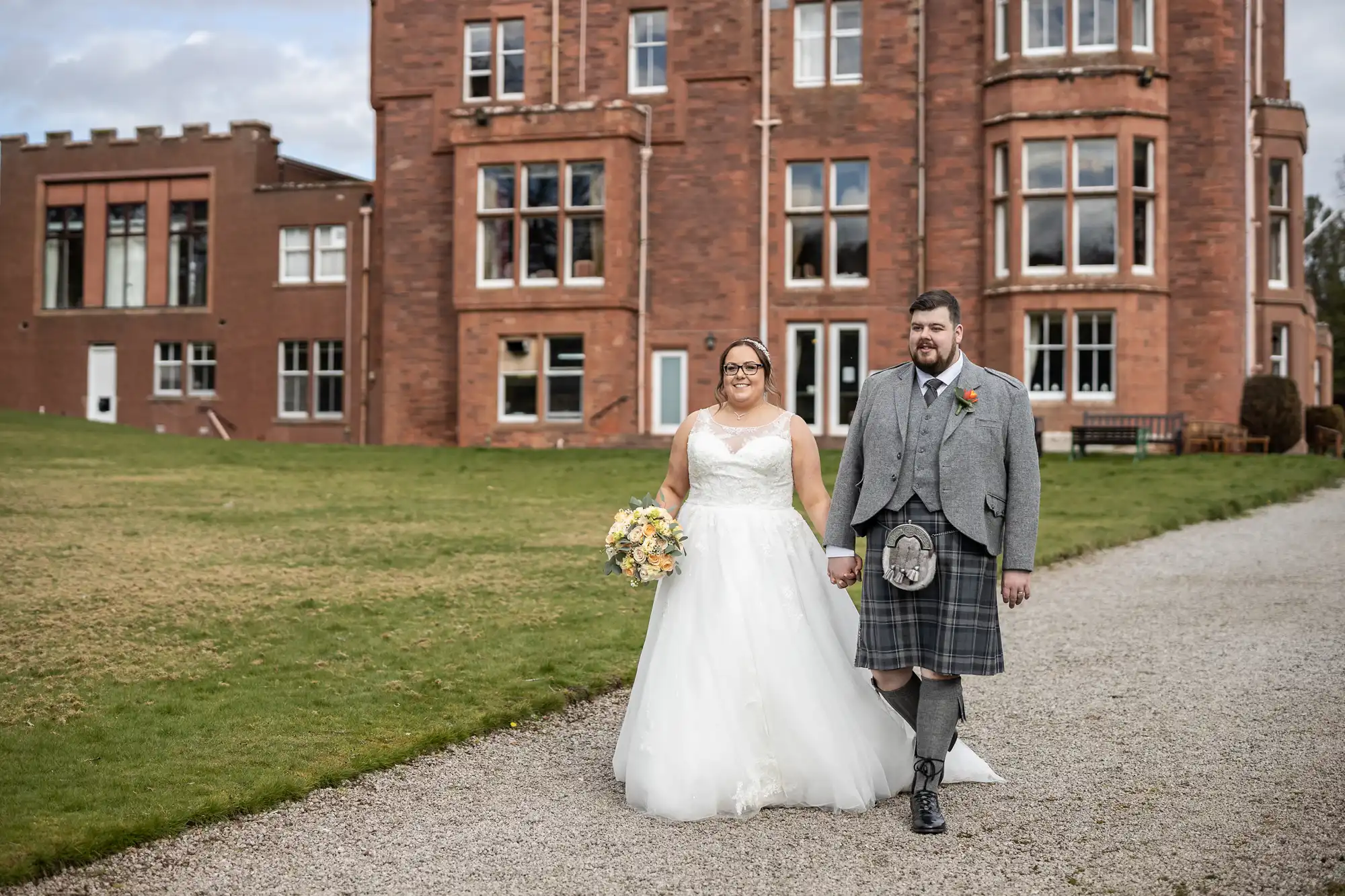 A bride in a white gown and a groom in a grey jacket and kilt walk hand-in-hand on a gravel path outside a large red-brick building with many windows, on a lawn under a partly cloudy sky. The bride holds a bouquet of flowers and both are smiling.