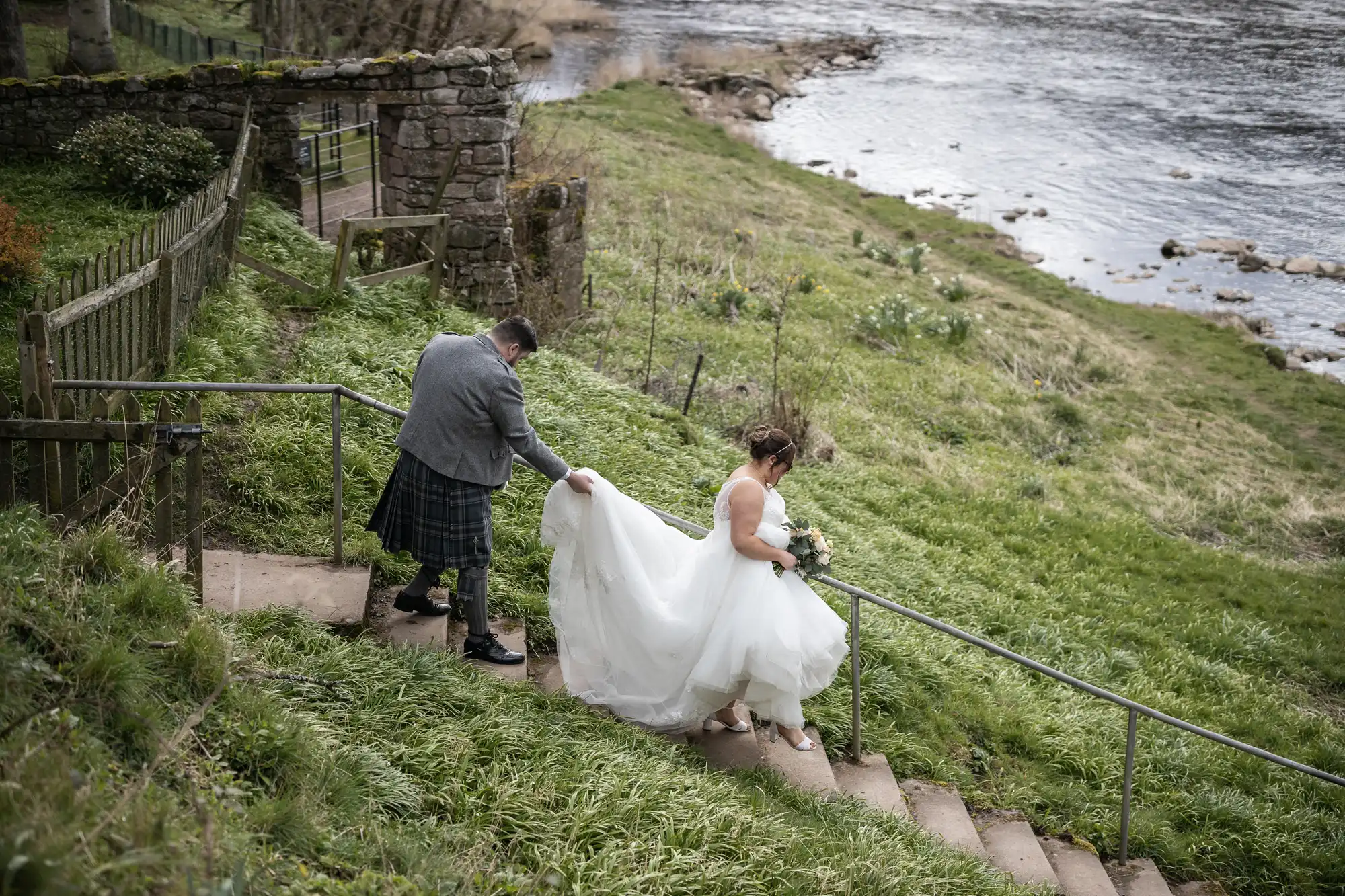 A bride in a white gown and a groom in a grey jacket and kilt walk up outdoor stone steps beside a grassy riverbank. The groom holds the bride’s dress train as she carries a bouquet, with a stone wall and river visible in the background.