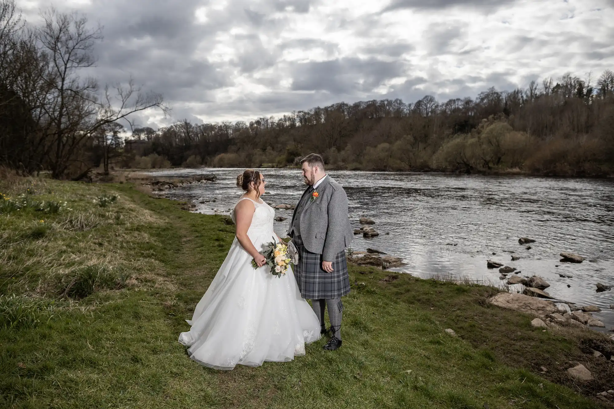 A bride in a white gown and a groom in a grey jacket and kilt stand on grass by a river, facing each other and smiling, with cloudy skies and a line of trees in the background. The bride holds a bouquet of yellow and white flowers.