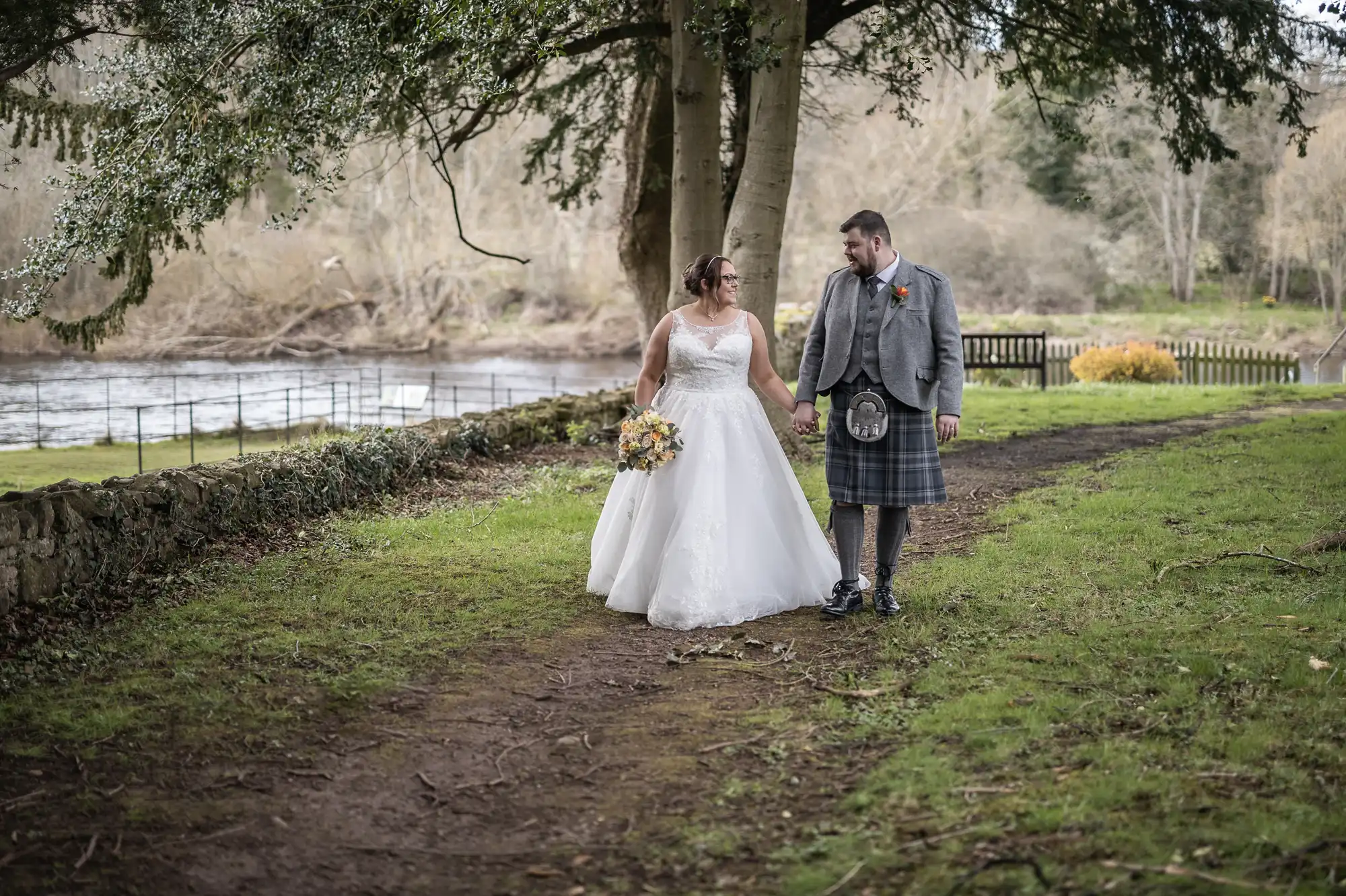 A bride in a white dress holding a bouquet walks hand-in-hand with a groom in a grey jacket and kilt. They stroll along a grassy, wooded path by a river, under large trees, smiling at each other. The background is peaceful and natural.