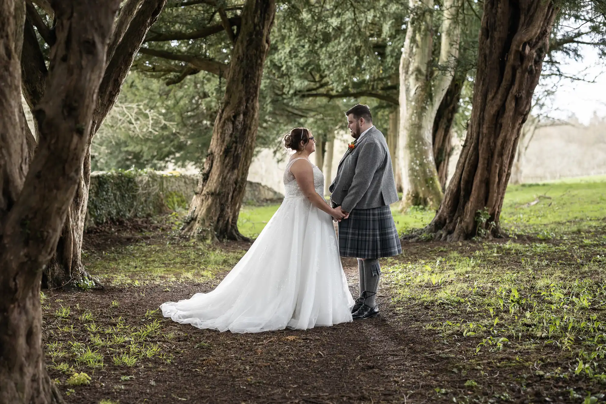 A bride in a white dress and a groom in a kilt hold hands, facing each other beneath tall, leafy trees in a sunlit, wooded area. The scene is serene with soft natural light filtering through the branches.
