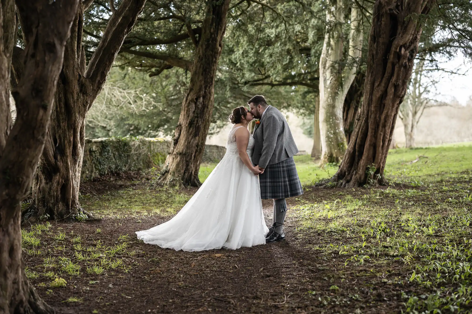 A bride in a white gown and a groom in a grey jacket and tartan kilt stand hand in hand, touching foreheads under tall trees with green grass and dappled sunlight, creating a romantic woodland wedding scene.