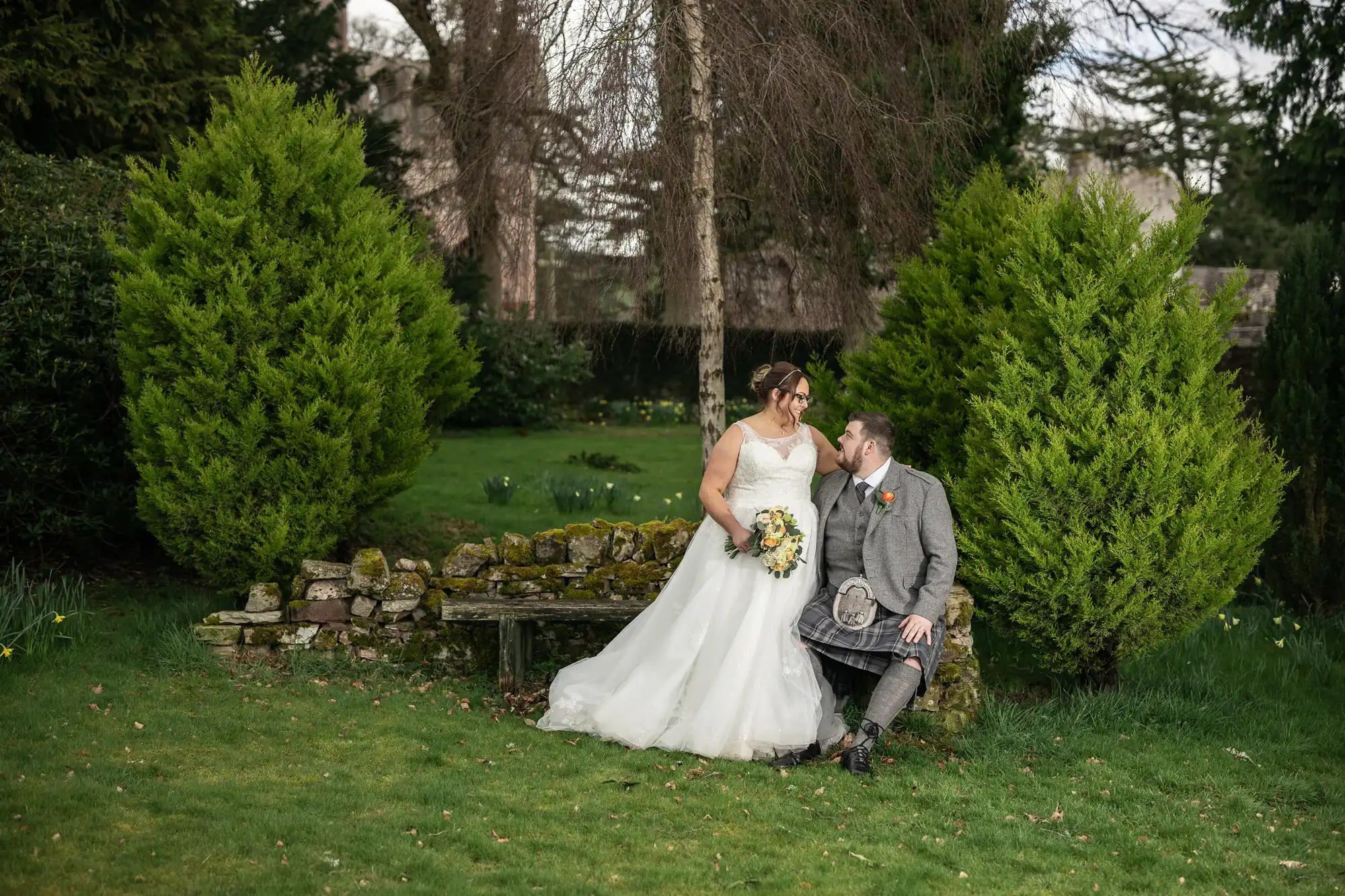 A bride in a white dress and a groom in a grey kilt outfit sit on a stone bench in a green garden, smiling at each other. The bride holds a bouquet, and they are framed by two tall, bushy evergreen trees.