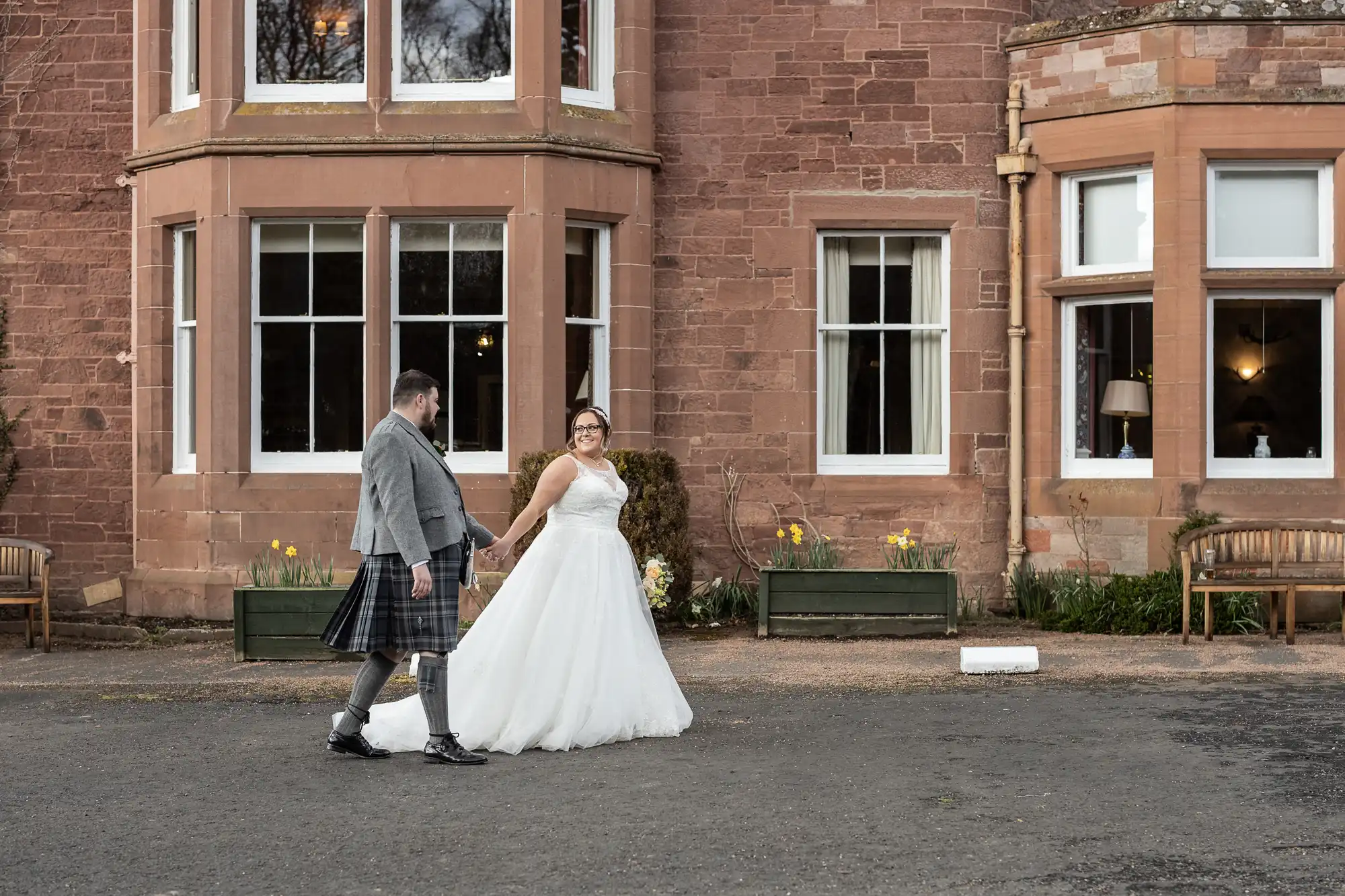 A bride in a white dress and a groom in a grey jacket and tartan kilt hold hands and smile as they walk outside a large brick building with white-framed windows and flower tubs. Benches and daffodils are visible in the background.