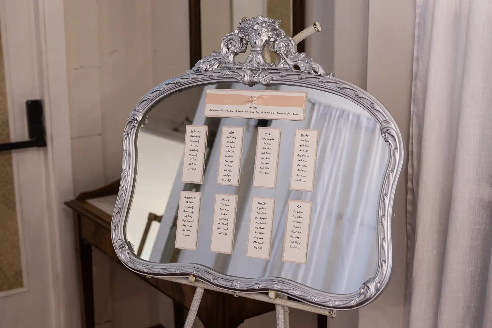 An ornate, vintage-style mirror on an easel displays a wedding seating plan. Lists of guest names are printed on white cards and arranged in columns on the mirror's surface. The background shows a wooden table and white curtains.