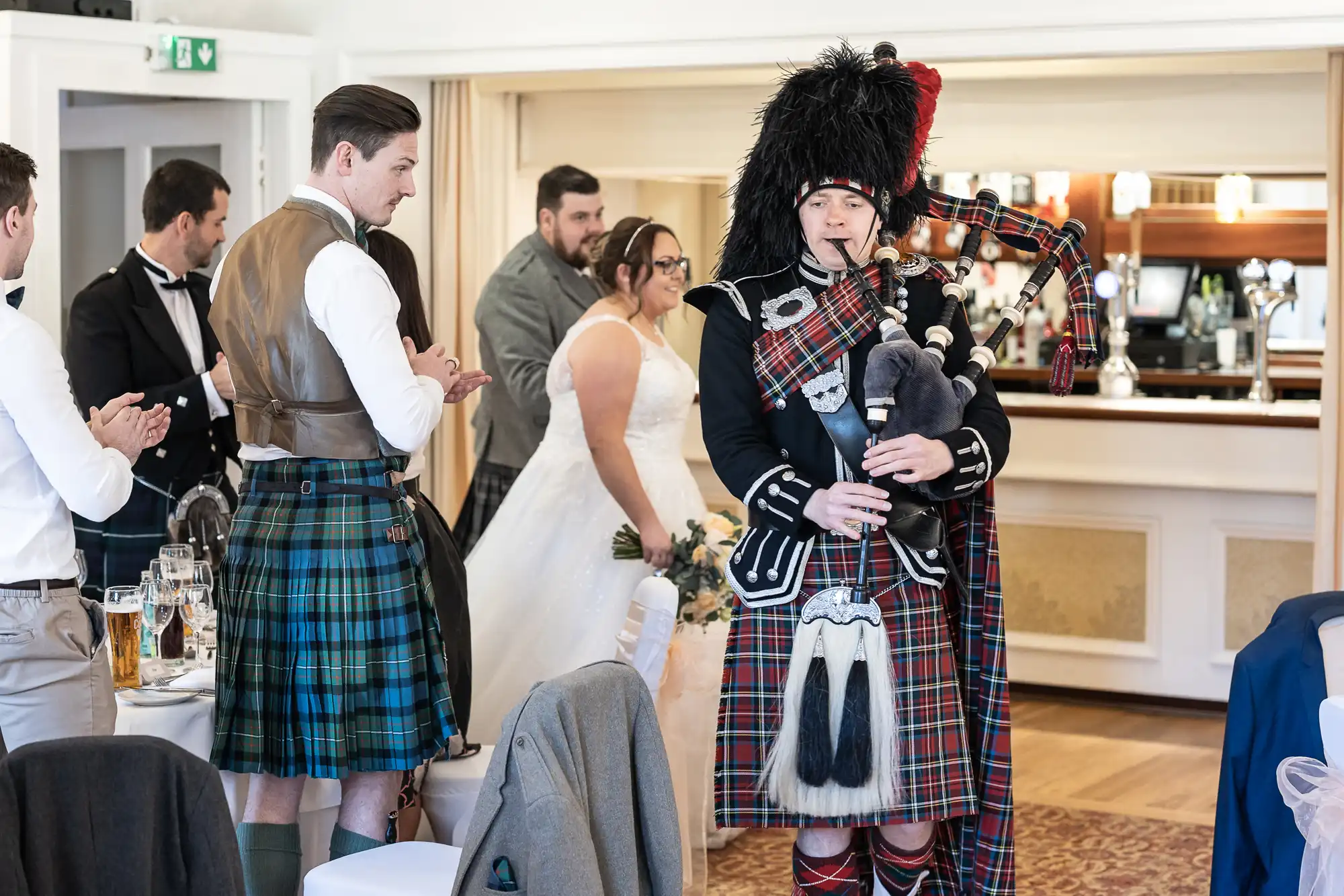 A bagpiper in traditional Scottish attire plays at an indoor wedding reception. Guests in kilts and formal wear stand and clap as a bride in a white dress walks past decorated tables, with a bar area visible in the background.