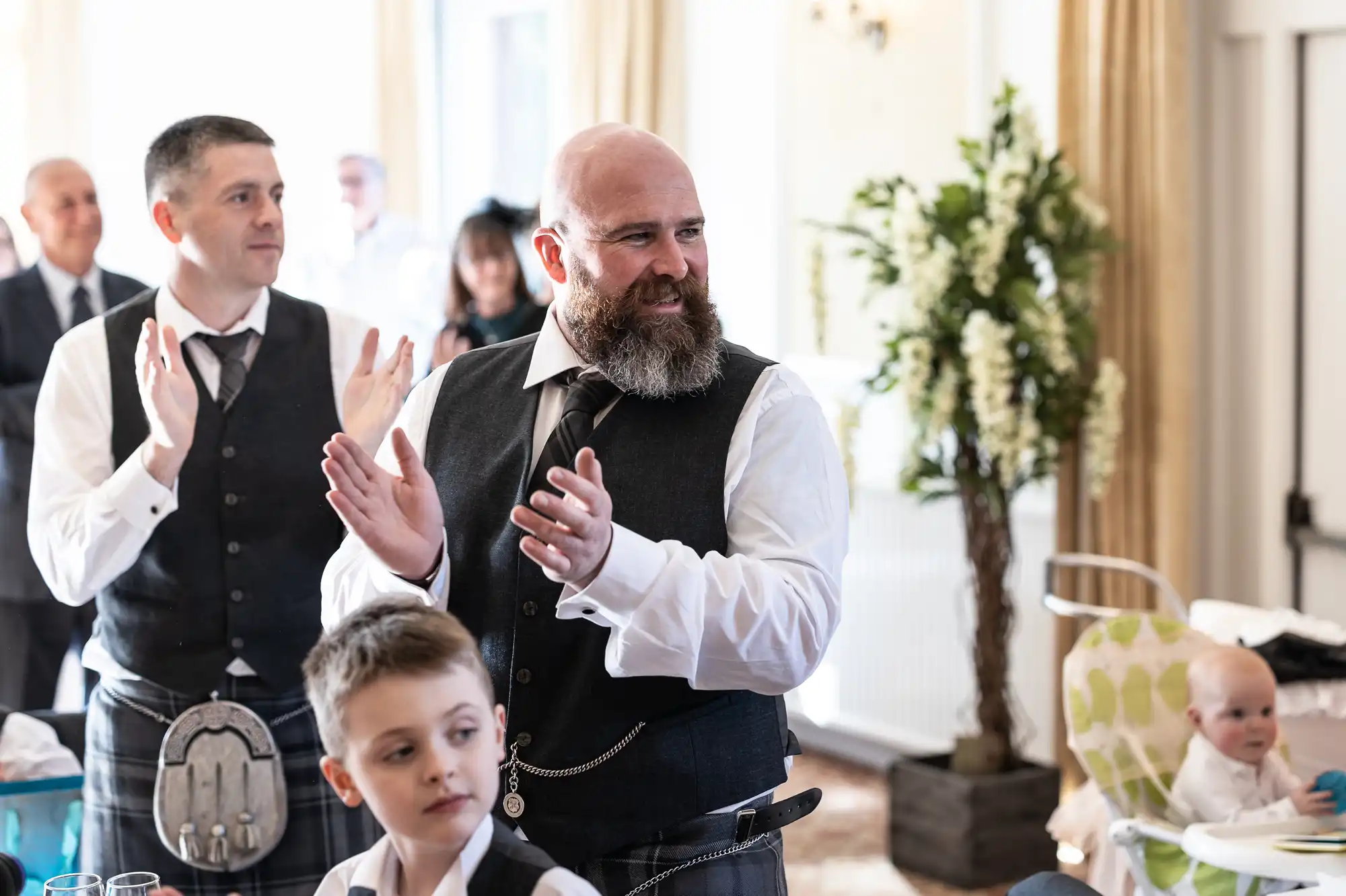 A bearded man in a black waistcoat and kilt claps at an indoor event, with another man in similar attire also applauding beside him. A young boy and a baby in a high chair are in the foreground. The room is bright with curtains and a large floral arrangement.
