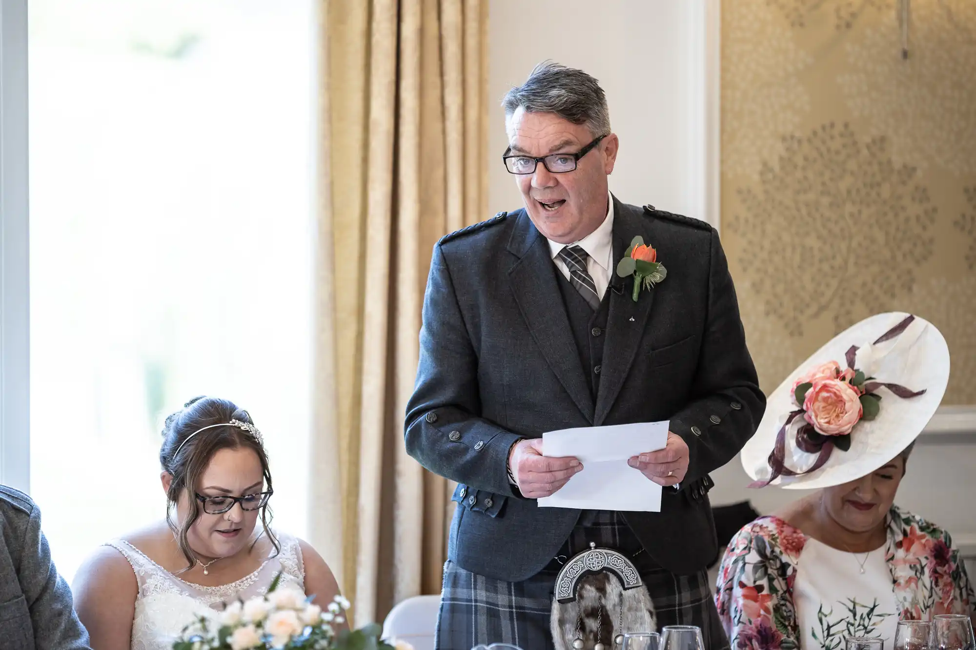 A man in a grey suit and checked kilt stands holding papers, speaking with an expressive face at a wedding reception. Seated beside him are a woman in a white wedding dress and another in a floral outfit with a large hat decorated with flowers.