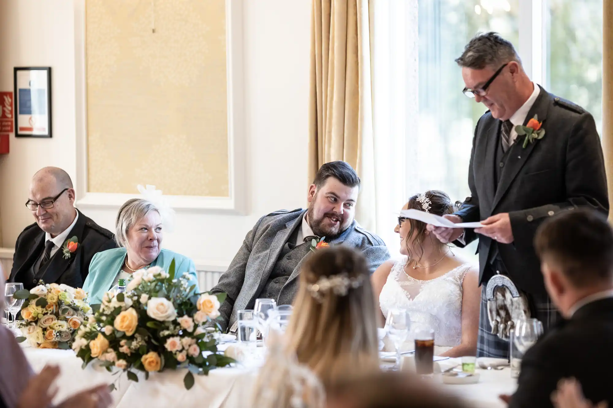 A wedding reception scene shows a groom smiling at his bride while a man in a suit reads from a paper. To their left, an older couple sits beside a floral centrepiece, all dressed formally. Guests are seated in the foreground, and sunlight filters through a window.