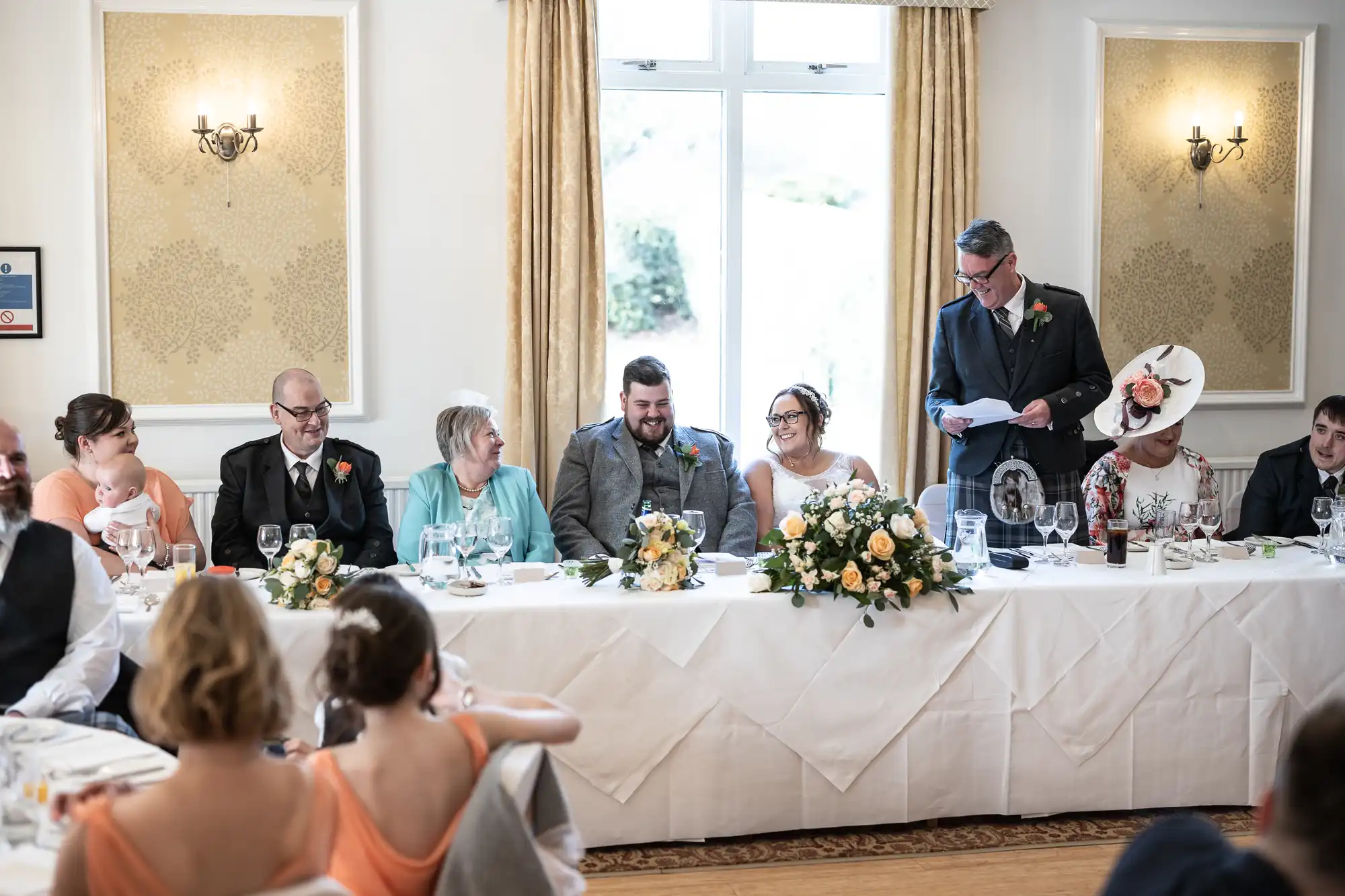 A wedding reception scene shows a long table with the bride and groom at the centre, surrounded by family and friends. A man in a suit stands giving a speech. Elegant floral arrangements and glasses decorate the table; guests listen and smile.