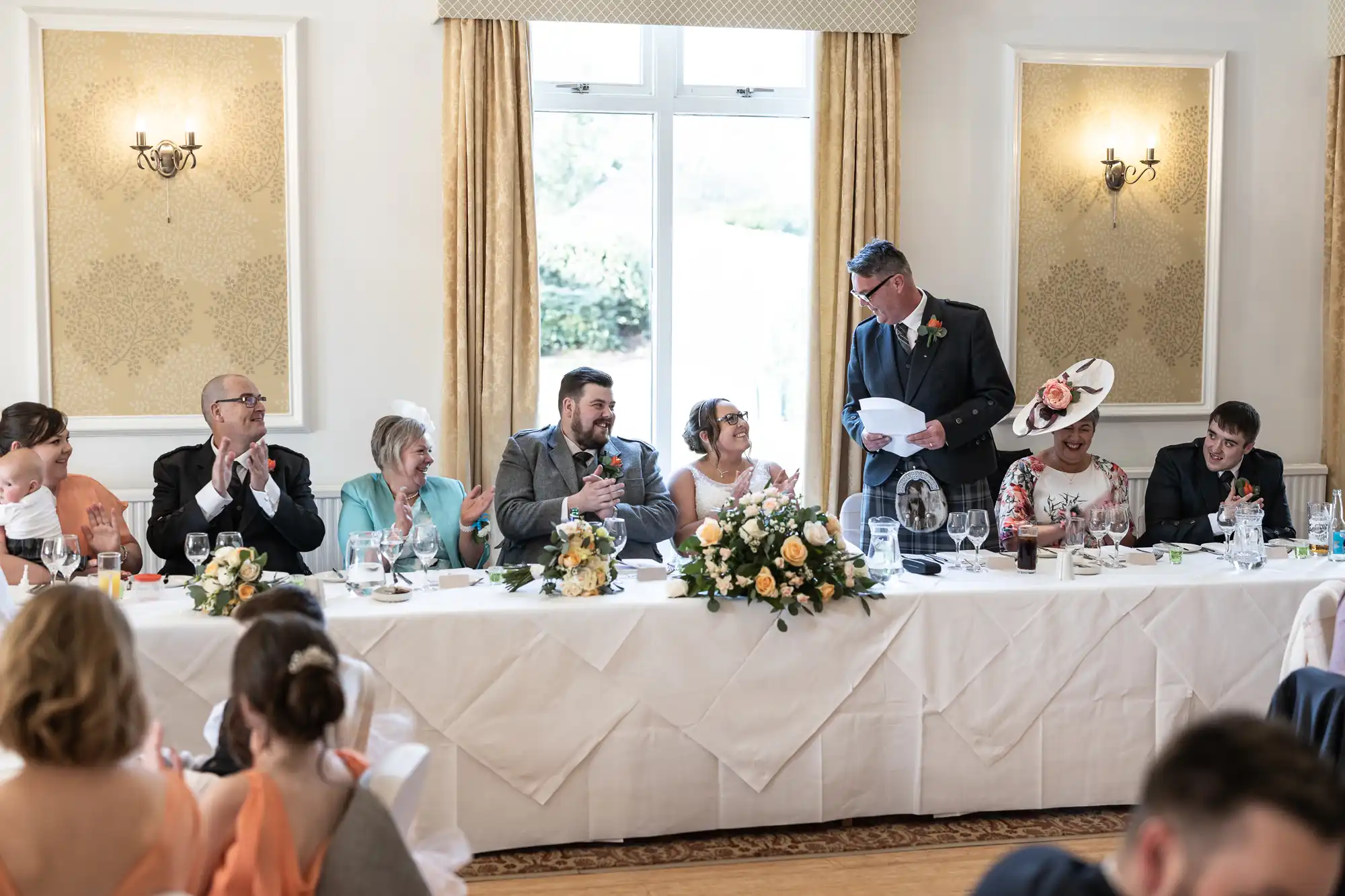 A wedding reception scene shows a bride and groom seated at a long table with family and friends. The groom and bride smile at a man standing and giving a speech. The table is decorated with flowers and glasses; guests applaud and listen.