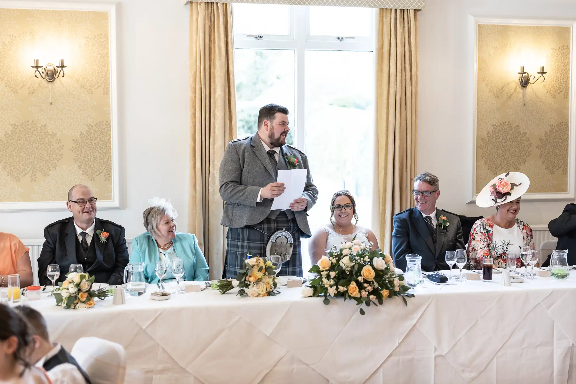 A man in a grey suit and tartan kilt stands giving a speech at a wedding reception table. Six others sit, smiling, with formal attire, flowers, and drinks in front of them, under bright windows and ornate gold wall decor.