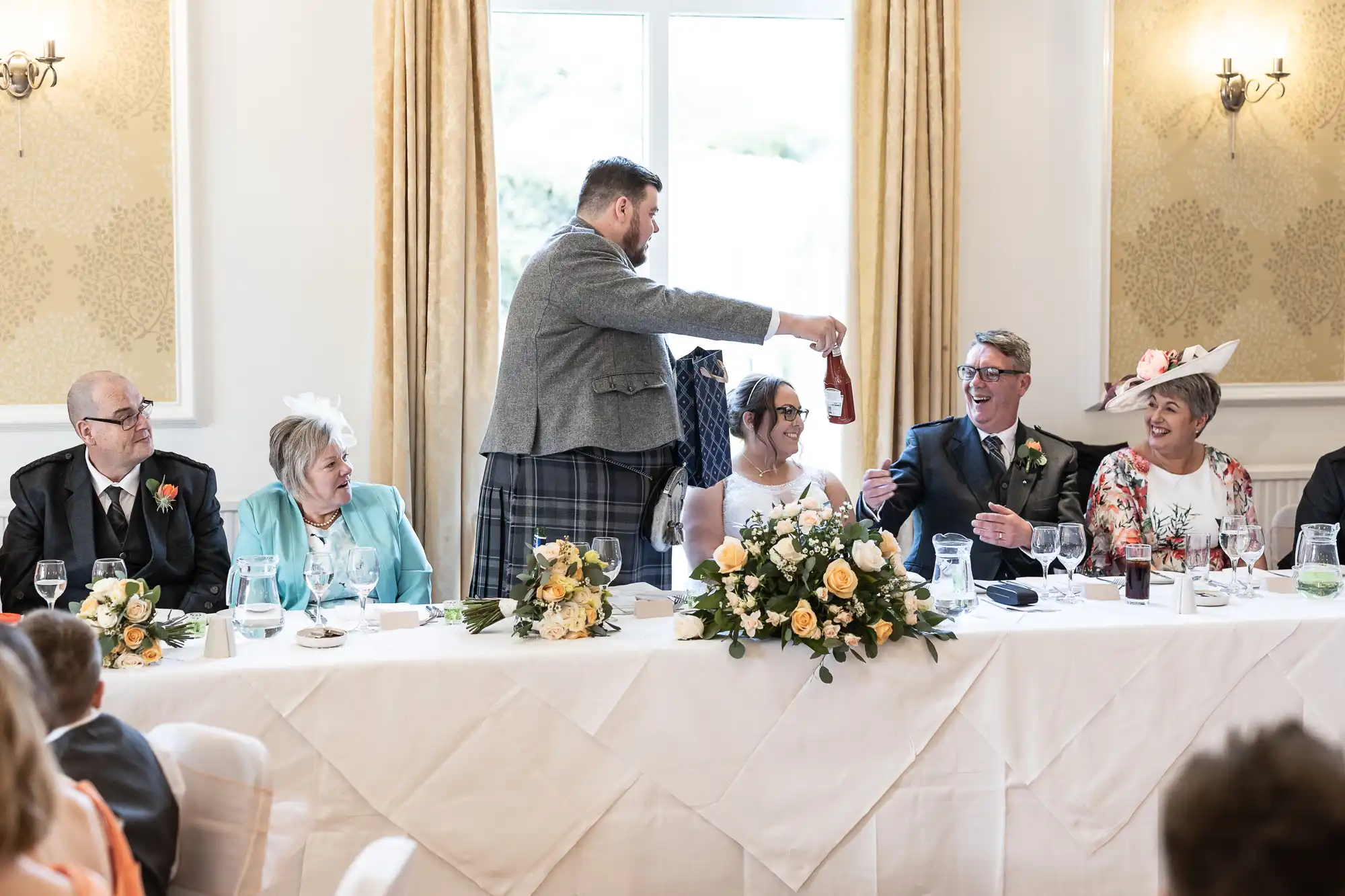 At a wedding reception, seven people sit at a long, decorated table. A man in a grey jacket stands, handing a wine bottle to a smiling man in a suit. The bride and others around them are laughing. Floral arrangements and glasses are on the white tablecloth.