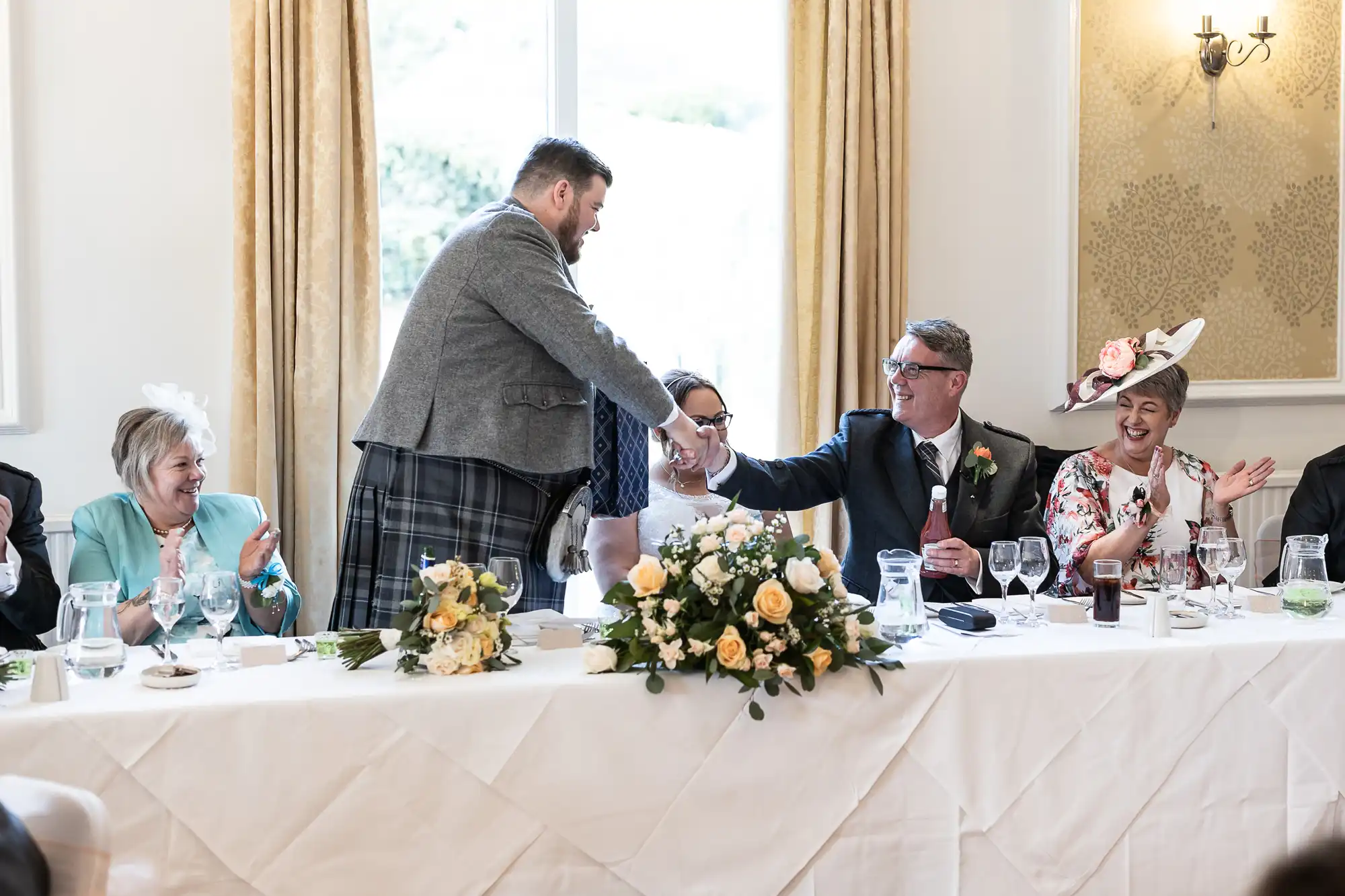 At a wedding reception, a man in a grey jacket and kilt shakes hands with a seated man in a suit. The bride, in white, sits between them, smiling. Other guests, including women in hats, sit at a decorated table with flowers and glasses.