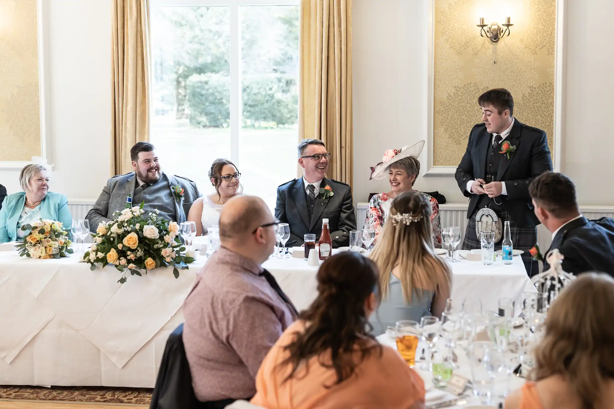 A wedding reception scene shows seven people seated at a long table, with one man standing and giving a speech. The group is smiling and laughing. The table is decorated with flowers and glasses. Other guests are seated in the foreground, facing the main table.