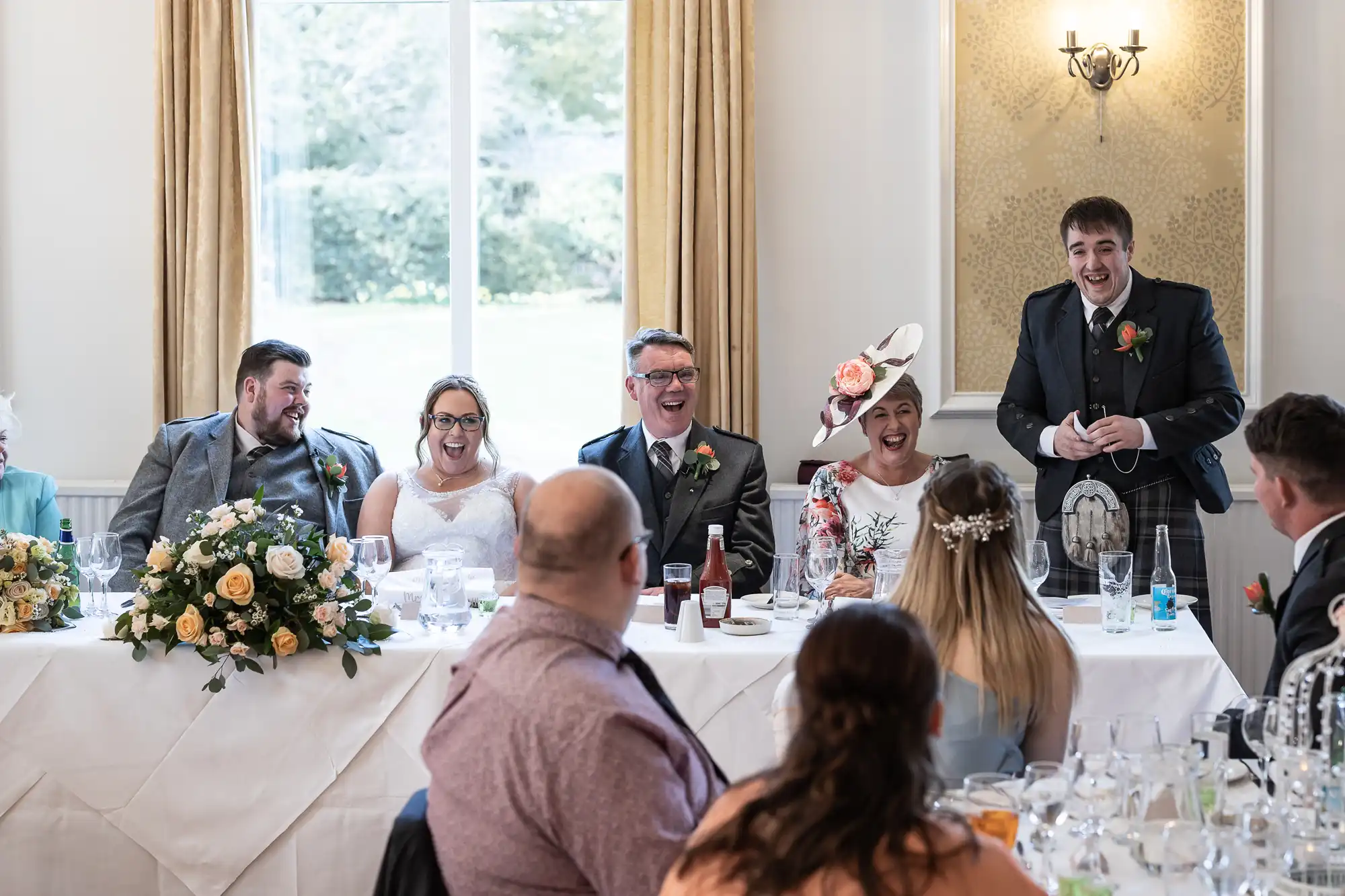 A wedding reception scene shows six people seated at a top table, laughing as a man in a suit stands giving a speech. The bride and groom are smiling, surrounded by family and flowers. Guests sit in the foreground, facing the speakers.