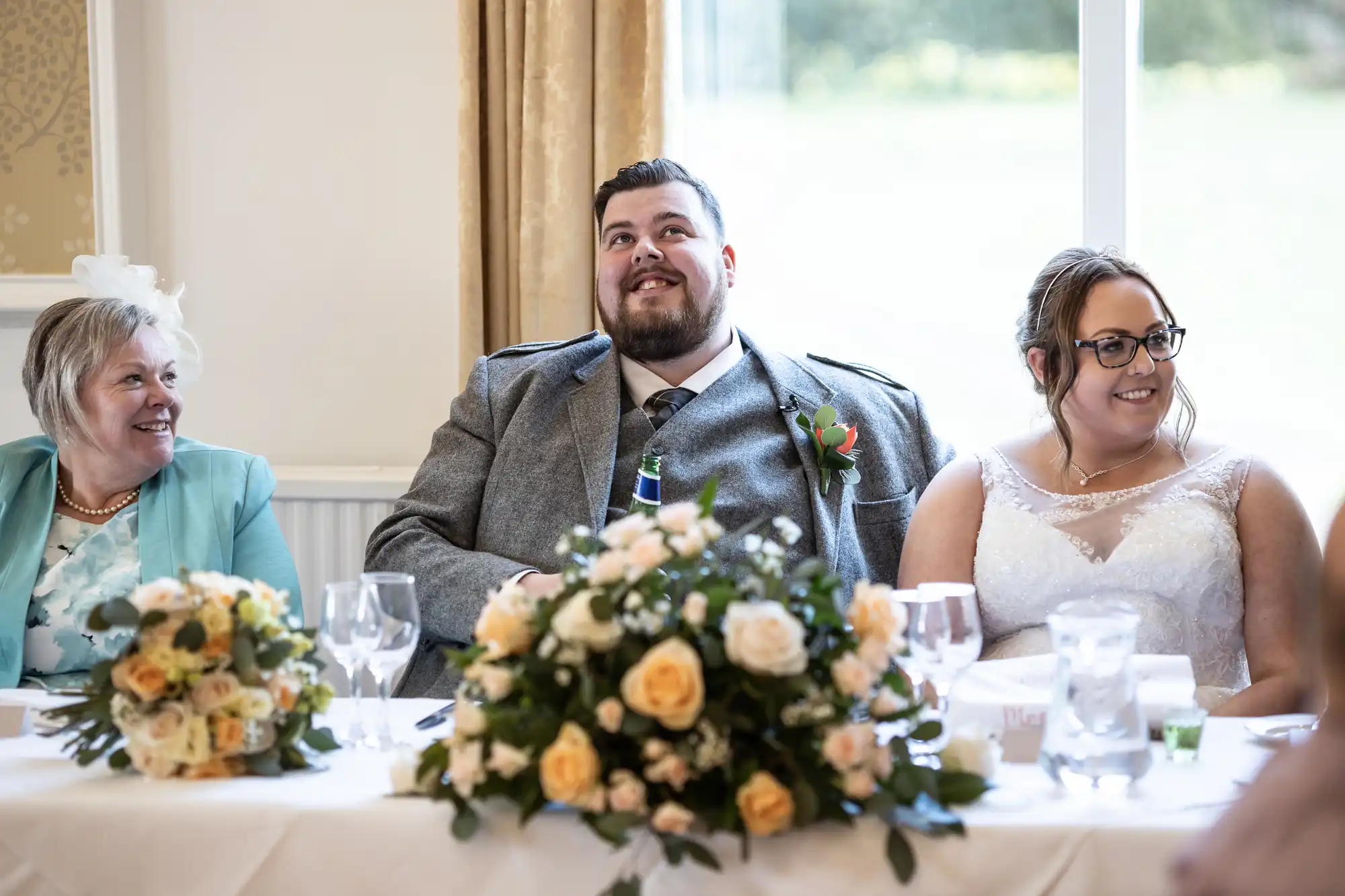 A joyful wedding reception scene: a bride in a white dress and glasses sits beside the groom in a grey suit, with an older woman in a light blue outfit. They sit behind a table adorned with glasses and a large yellow and white floral bouquet.