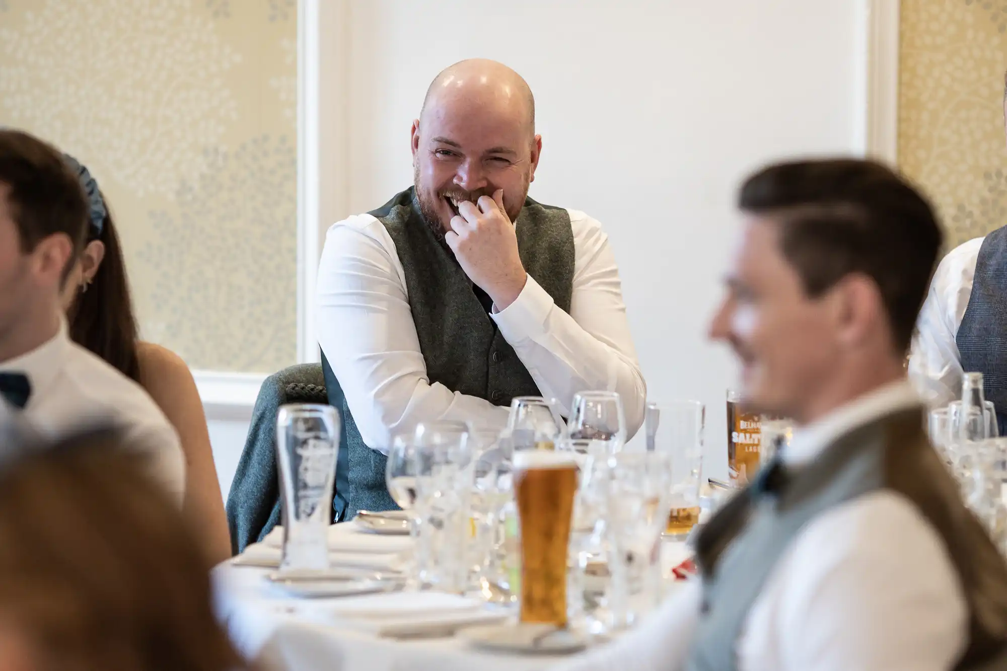 A bald man with a beard, wearing a white shirt and grey waistcoat, smiles with his hand near his mouth while seated at a table set with glasses and drinks. Other people in formal attire sit around him, all appearing to enjoy a light-hearted moment.