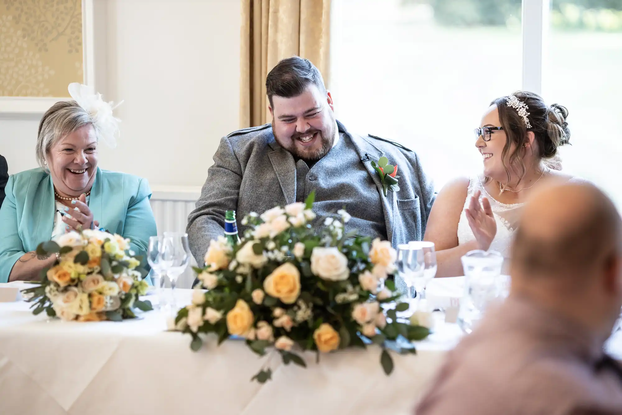 A smiling bride and groom sit at a decorated table with flower arrangements and glasses, clapping and laughing with a woman in a pale blue outfit and white hat, during a wedding celebration by a bright window.