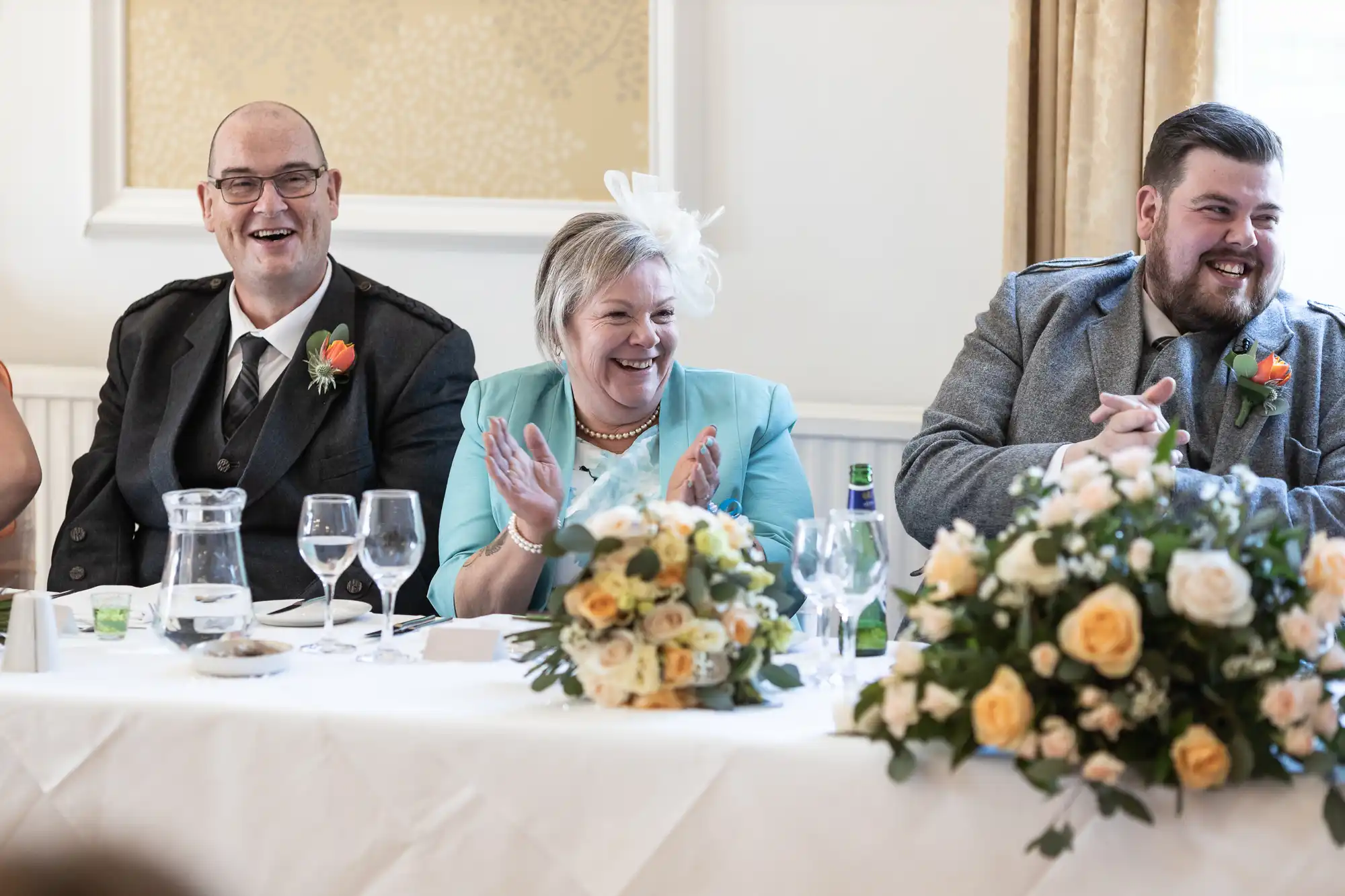 Three adults dressed formally sit at a wedding reception table decorated with yellow and white flowers. The woman in the middle claps and smiles, whilst the two men on either side laugh. Glassware and floral arrangements are on the white tablecloth.