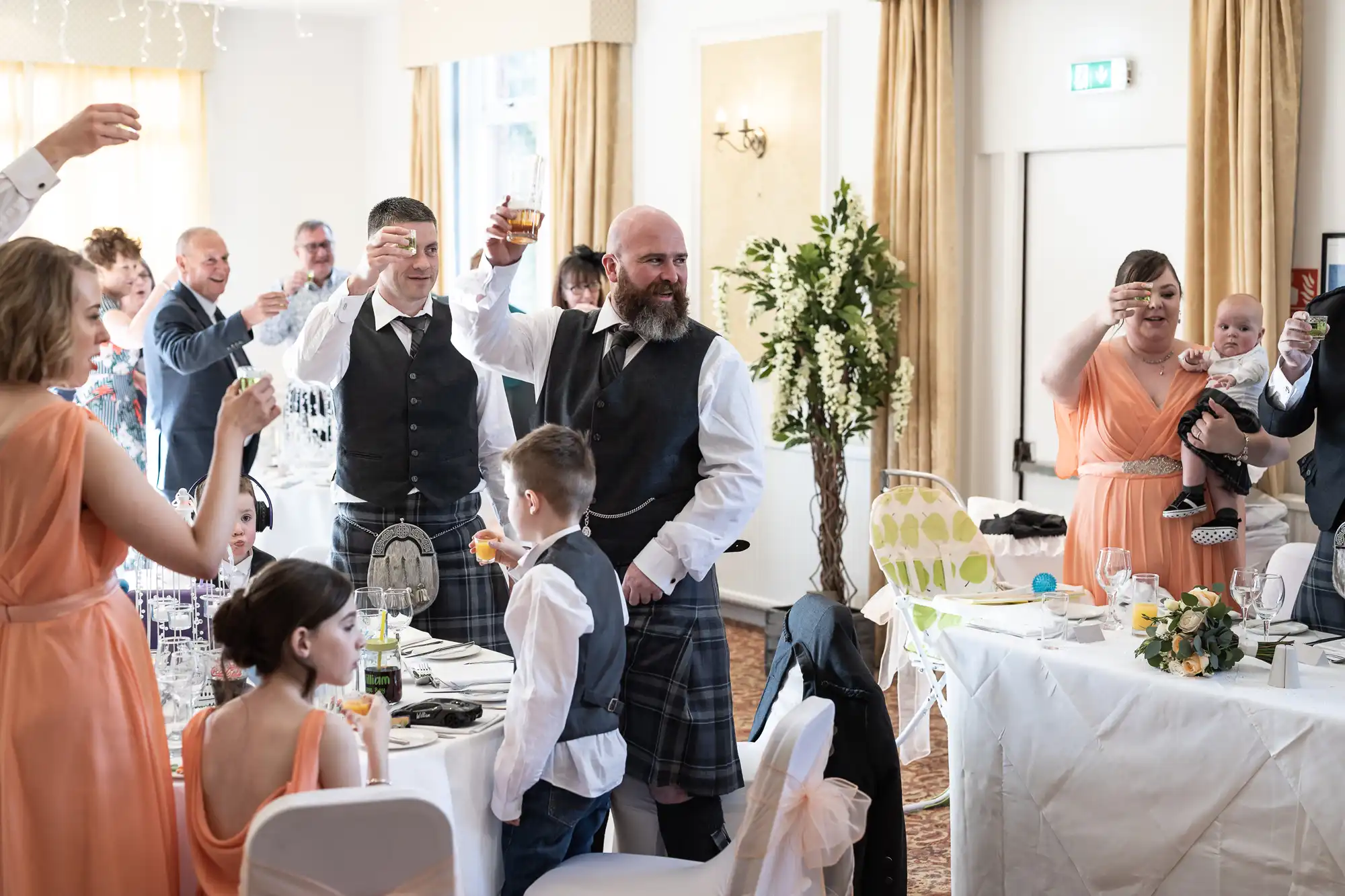 Guests at a wedding, some in kilts and waistcoats, stand around decorated tables raising glasses for a toast. Women in peach dresses, children, and a baby are present. Sunlight streams in through large windows, and the room is adorned with flowers and white linen.
