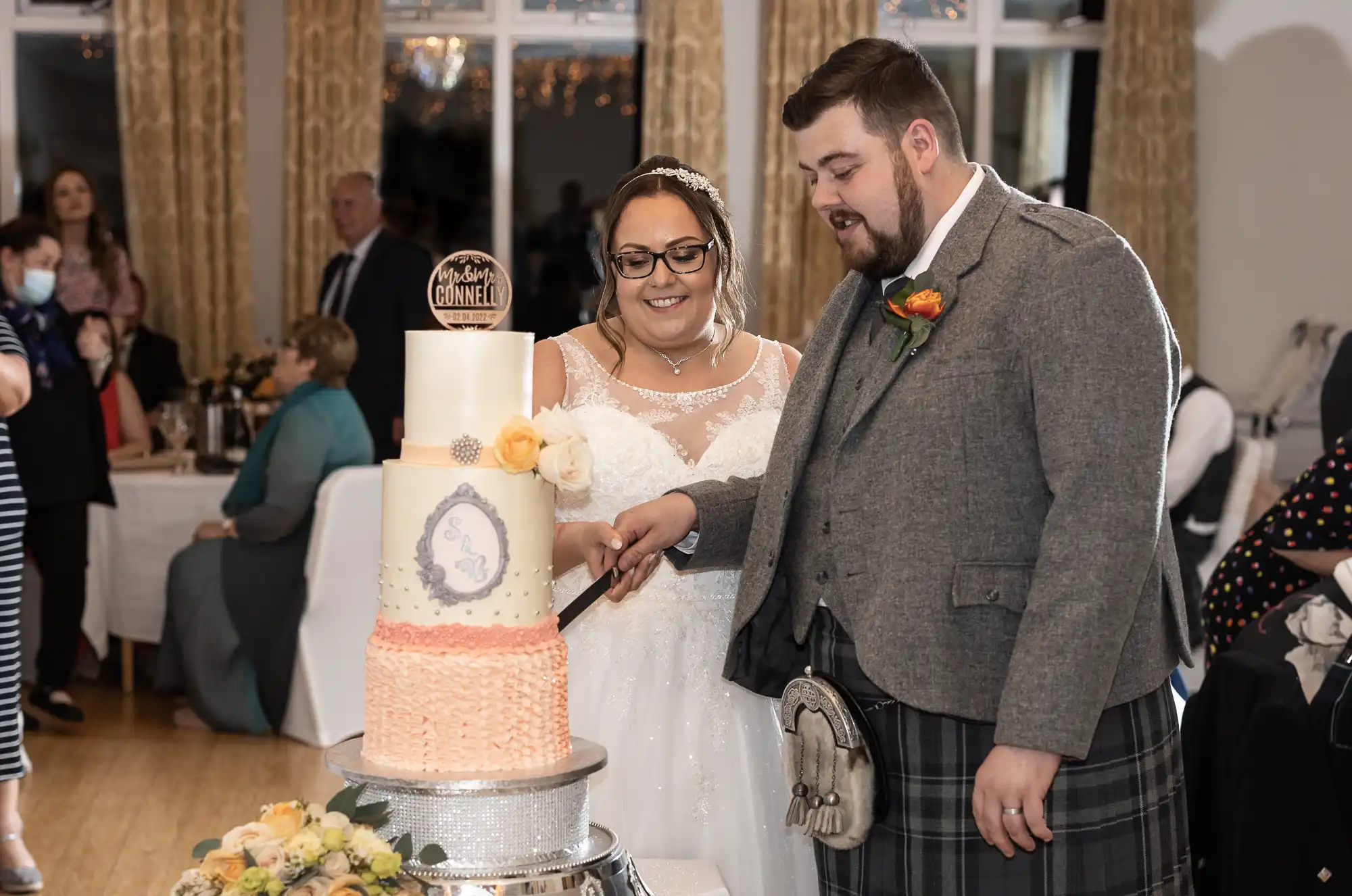 A bride in a white dress and a groom in a grey jacket and kilt cut a three-tier wedding cake together. The cake is decorated with flowers and a topper. Guests are seated at tables in the background, and the room has tall curtains and bright lighting.