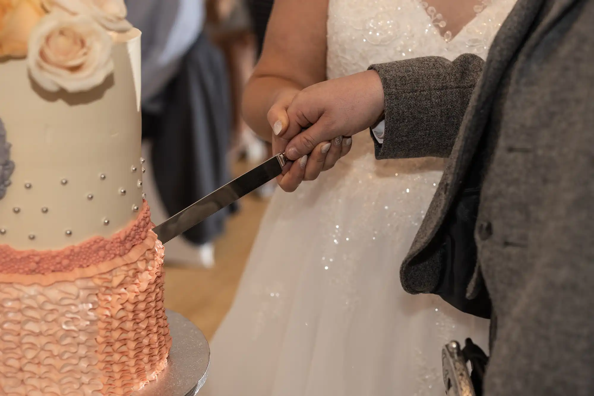 Close-up of a bride in a white wedding dress and a groom in a grey suit cutting a tiered wedding cake together. Both hold the knife, and the cake is decorated with textured pink icing and pastel flowers on top.