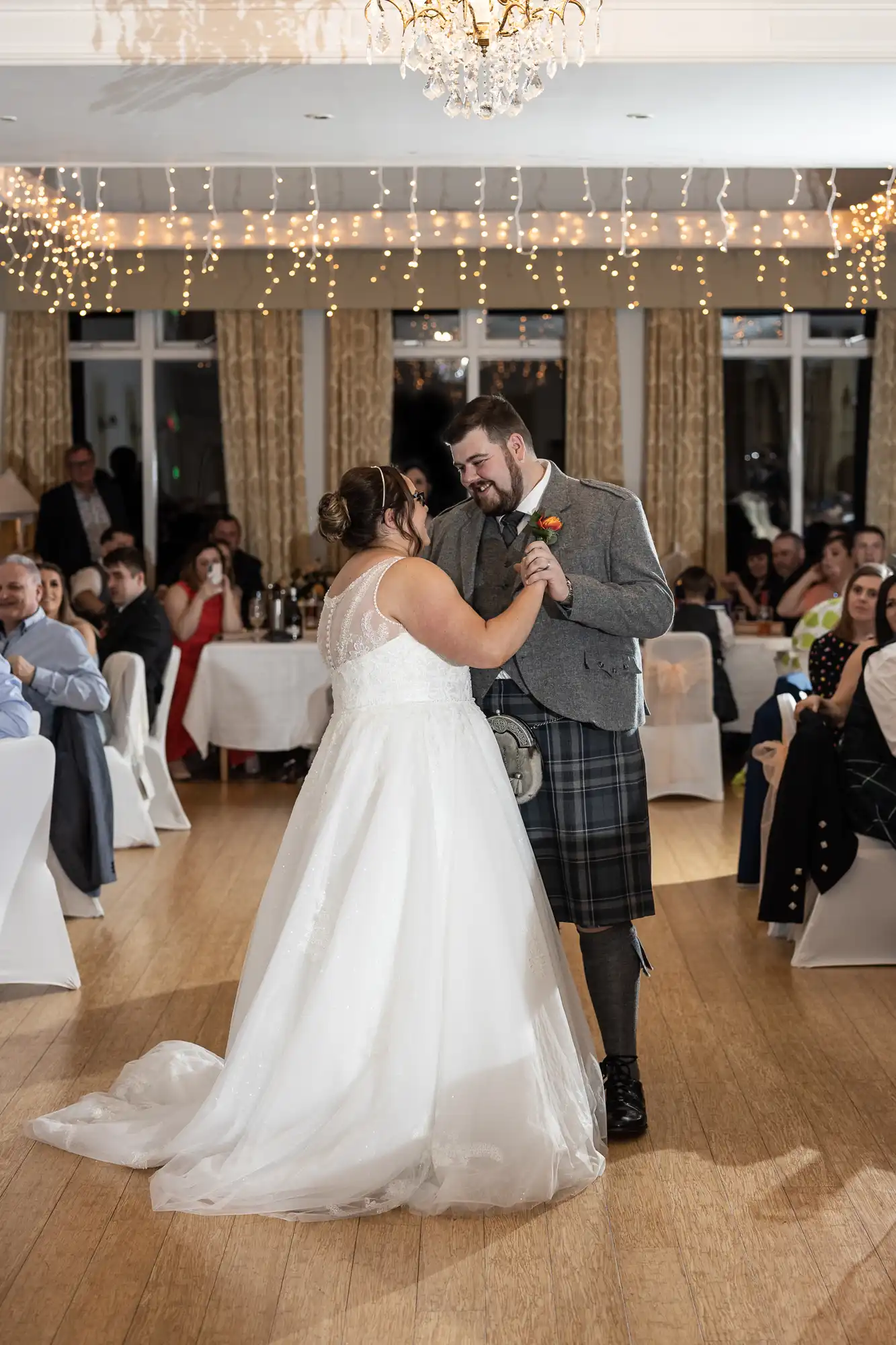 A bride in a white gown and groom in a grey jacket with a kilt share a first dance in a warmly lit reception hall. Guests seated around round tables watch them. Fairy lights and elegant curtains decorate the room.