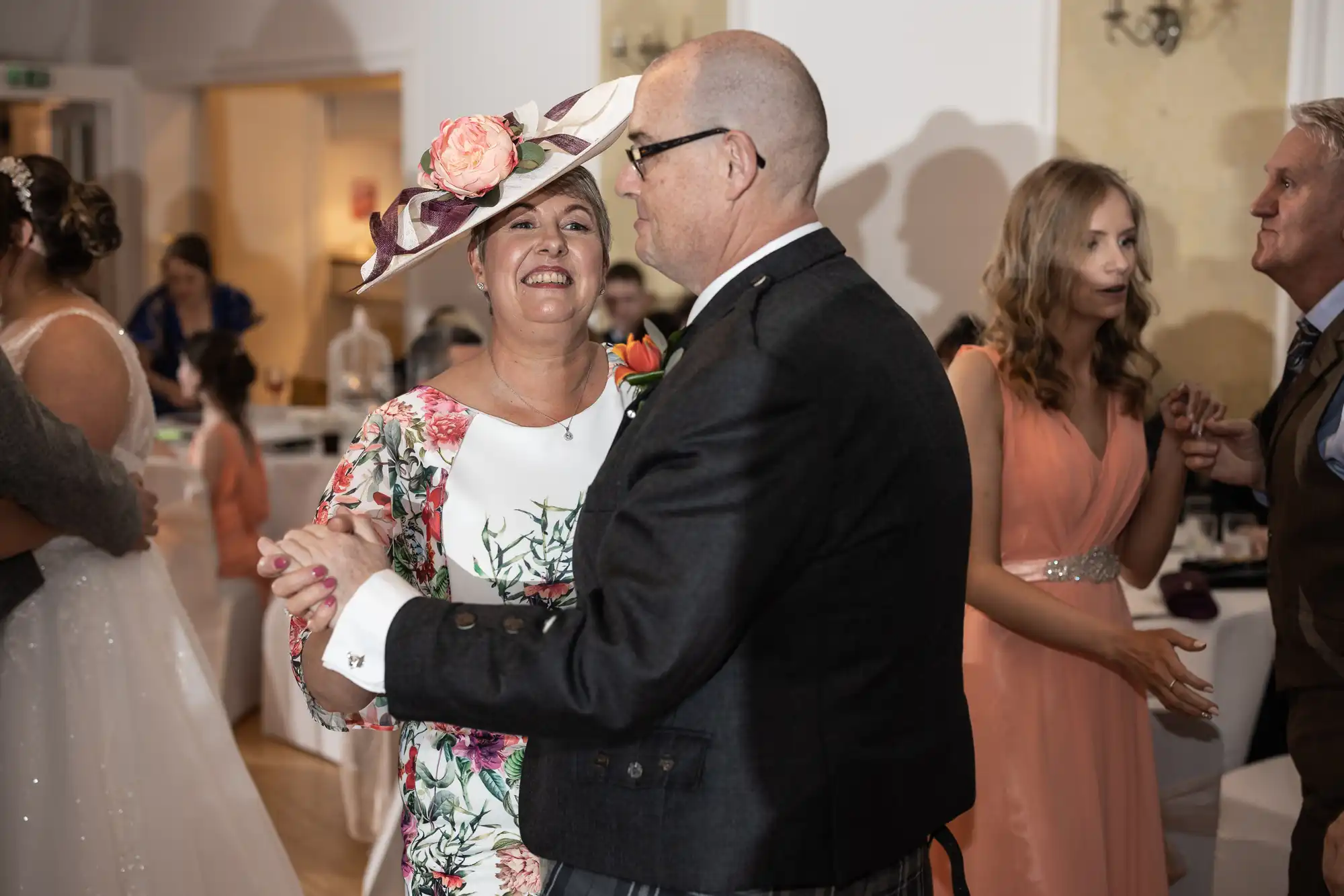 A smiling woman in a floral dress and large hat dances with a man in a dark suit at a wedding reception. Other guests in formal attire, including a woman in a peach dress, mingle and dance in the softly lit room.