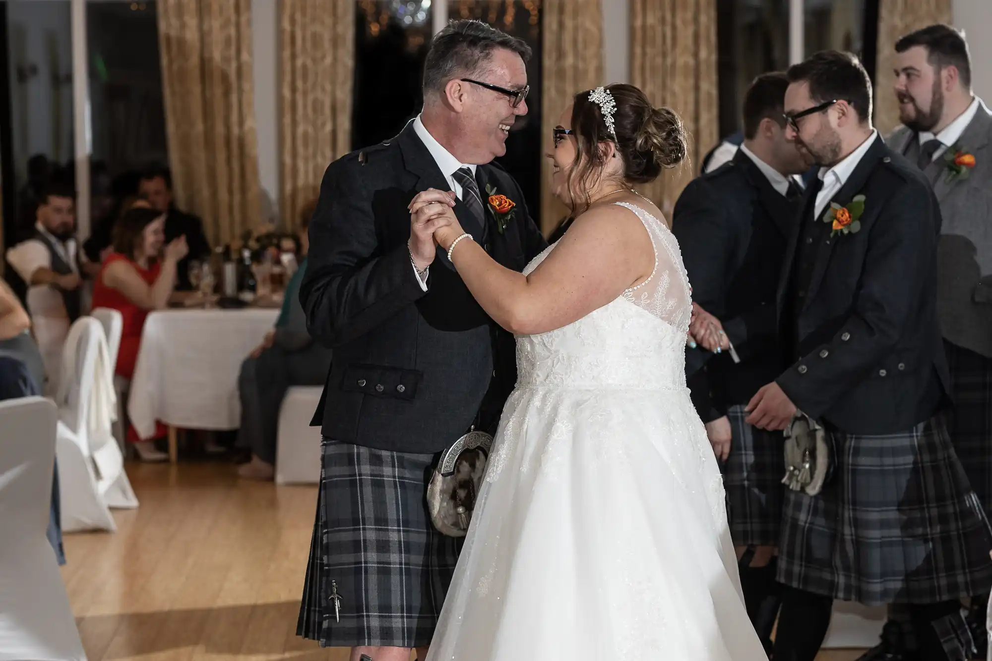 A bride in a white gown and a man in a dark jacket and tartan kilt dance together, smiling. Men in kilts and dark jackets stand nearby. Seated guests watch in a warmly lit room with golden curtains and white-draped tables.