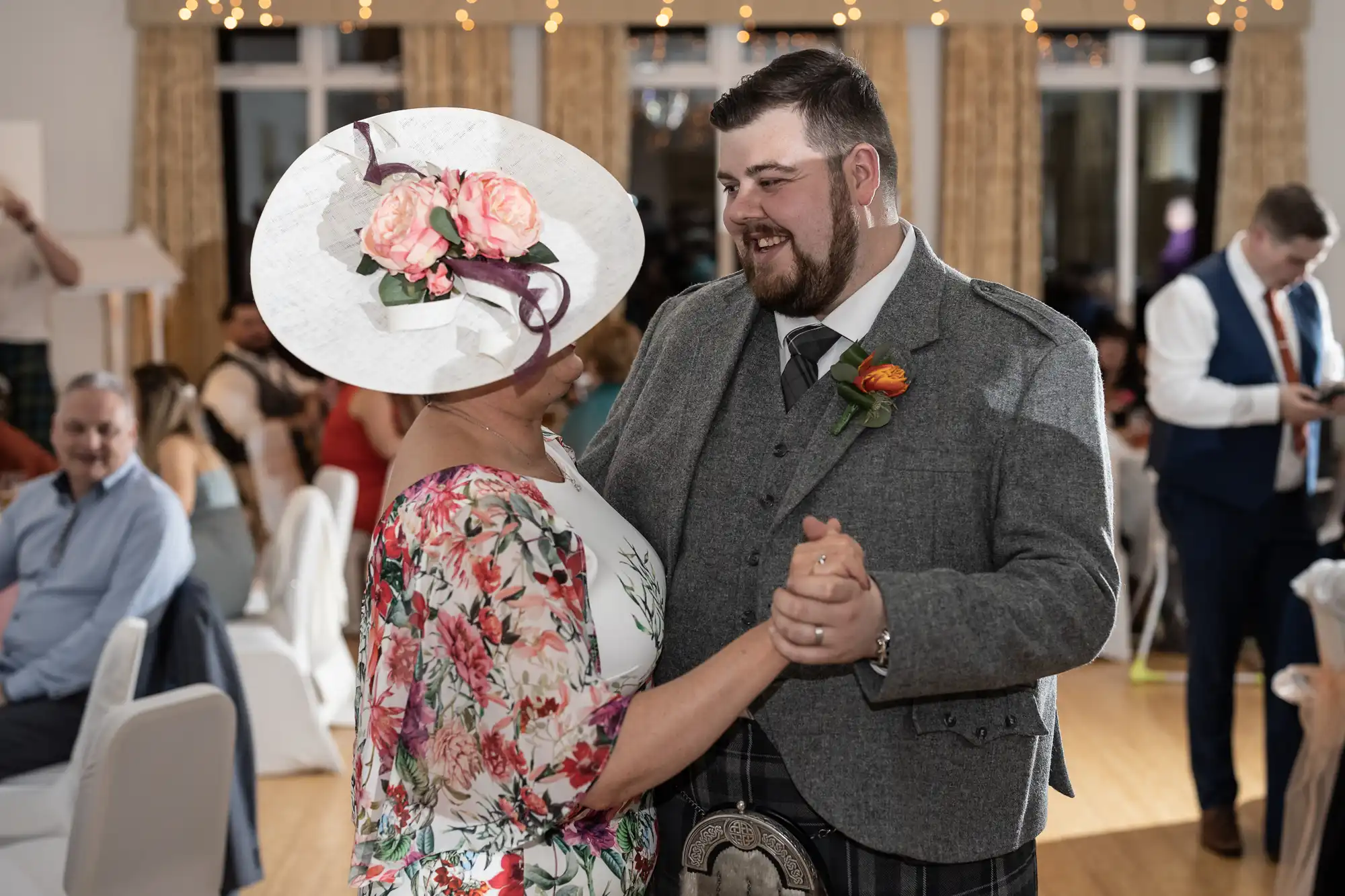 A man in a grey suit and kilt dances with a woman wearing a floral dress and large white hat with pink flowers. They smile at each other in a bright, decorated room with guests seated and mingling in the background.