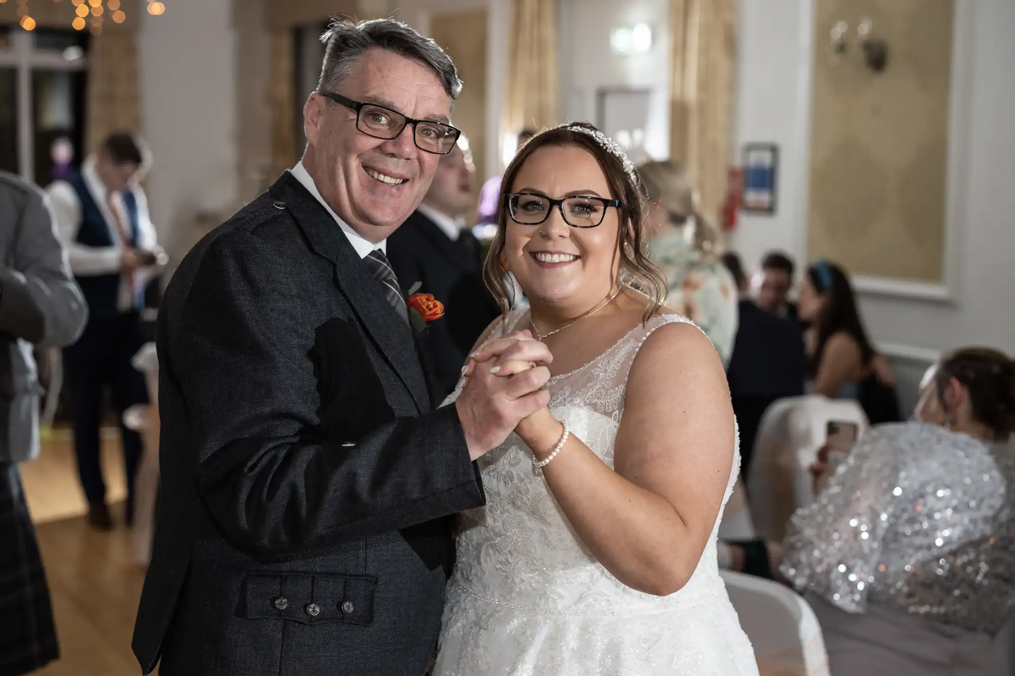 A smiling man in a dark suit and a woman in a white wedding dress hold hands and dance at a brightly lit indoor event, likely a wedding reception. Other guests are seated and mingling in the background. Both look happy and are facing the camera.
