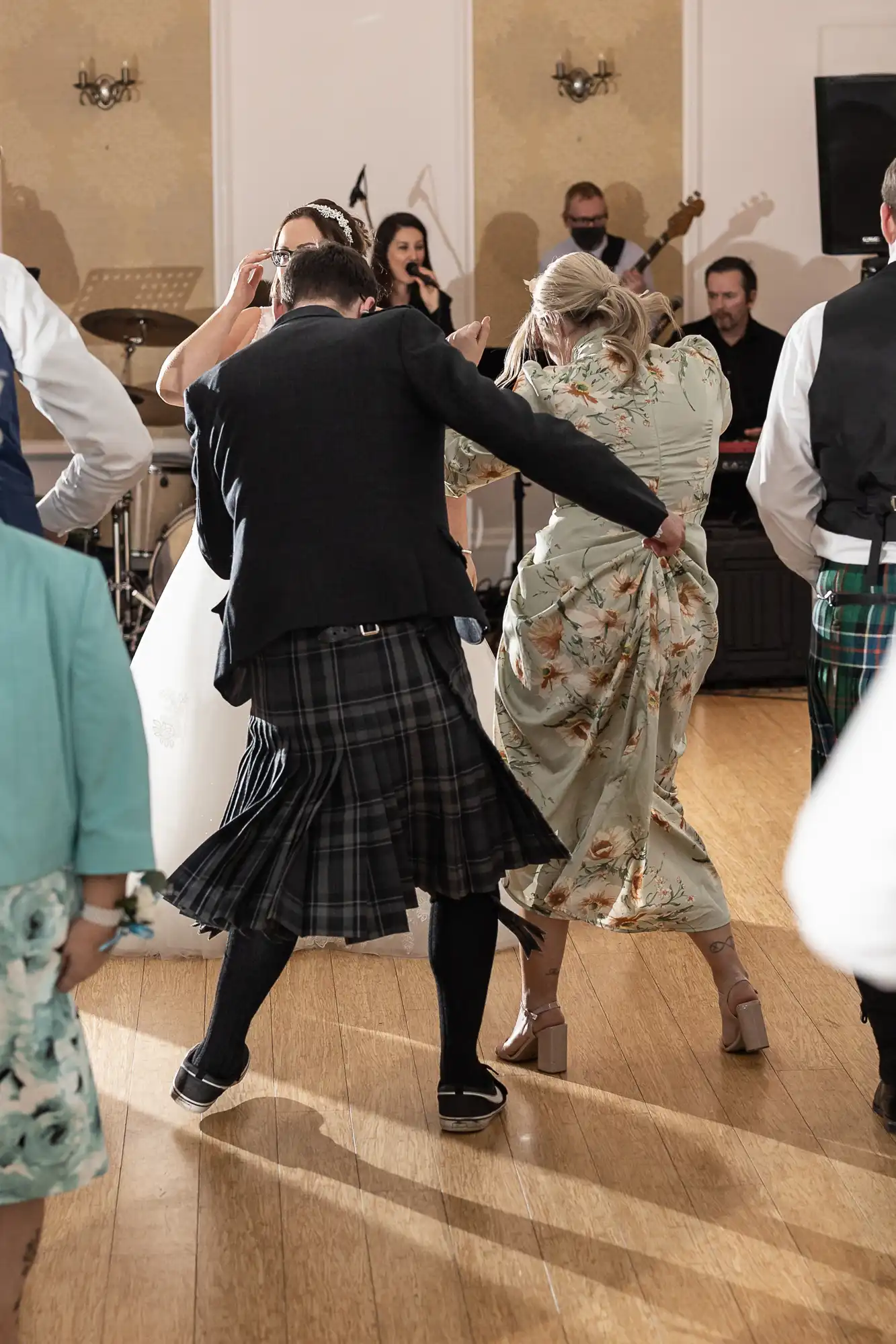 A man in a dark jacket, kilt, and knee-high socks dances energetically with a woman in a long floral dress at a lively indoor event. Other guests and a band with instruments are visible in the background. Light streams in, highlighting the dancers.
