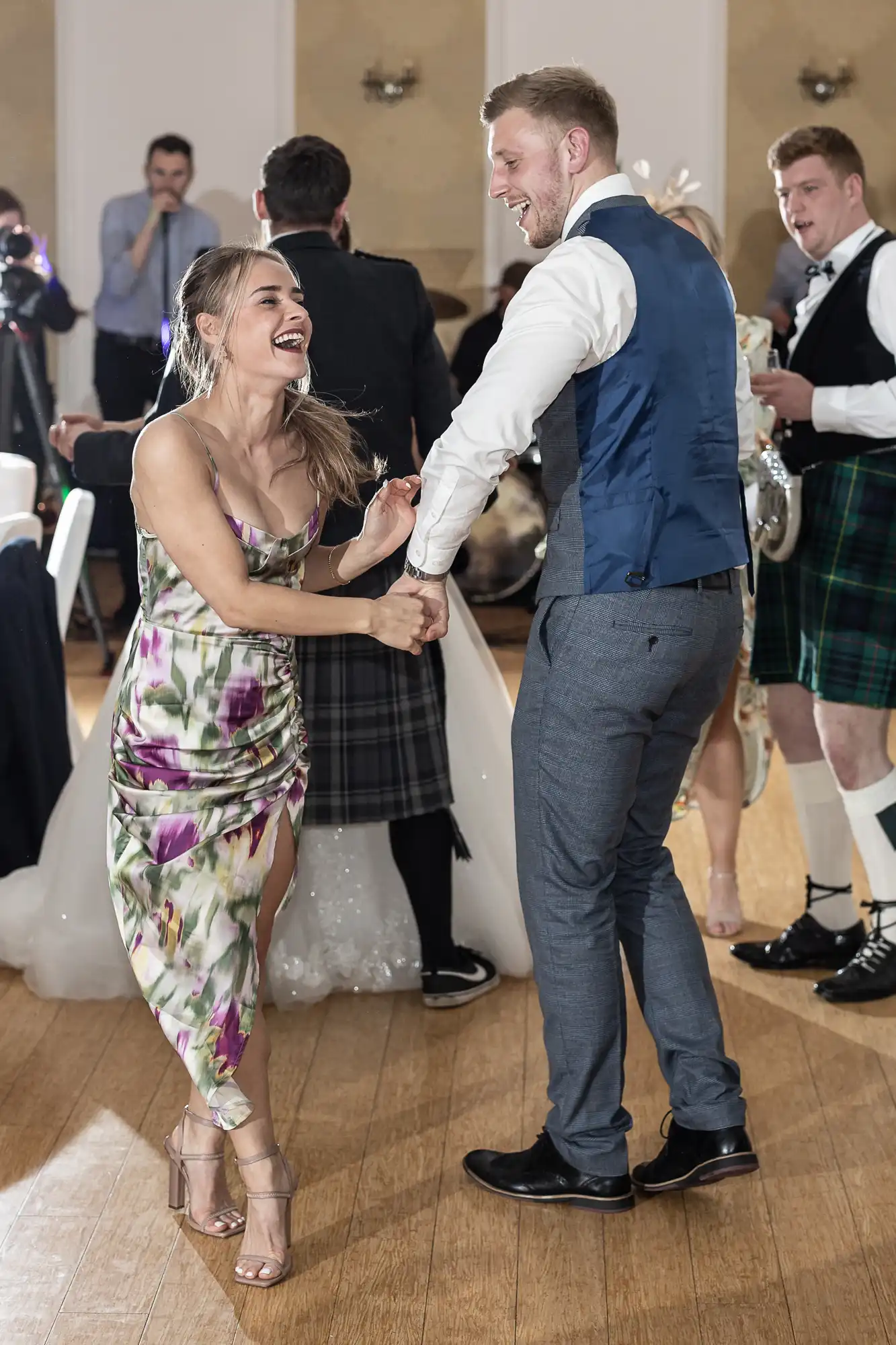 A smiling woman in a floral dress and a man in a blue waistcoat and tie dance together on a wooden floor at a formal event. People in kilts and evening attire, including a bride in a white dress, are seen celebrating in the background.