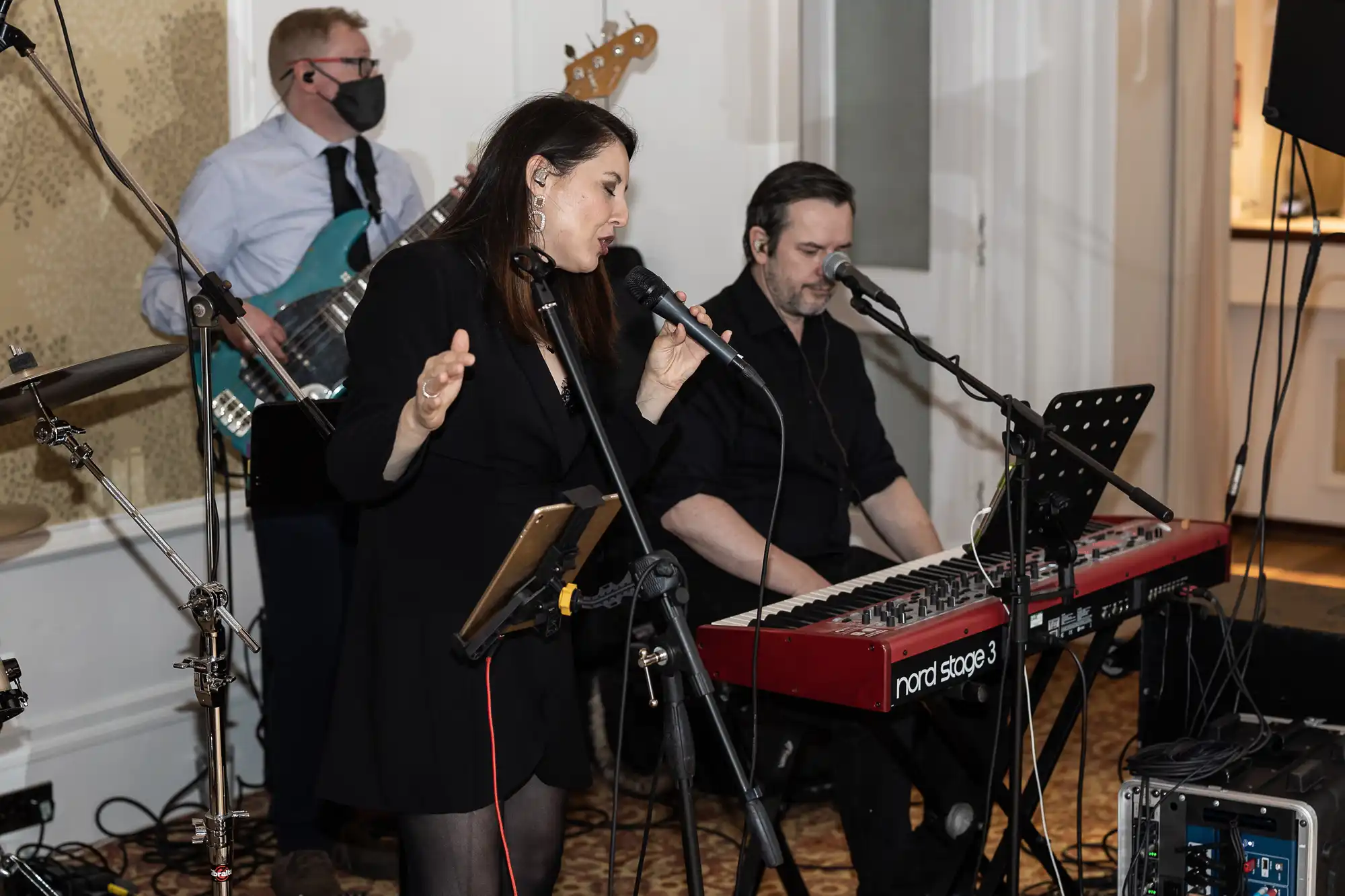A woman sings into a microphone while a man plays a keyboard beside her. Another man in the background plays bass guitar and wears a face mask. Music equipment and microphones are set up around them in an indoor venue.